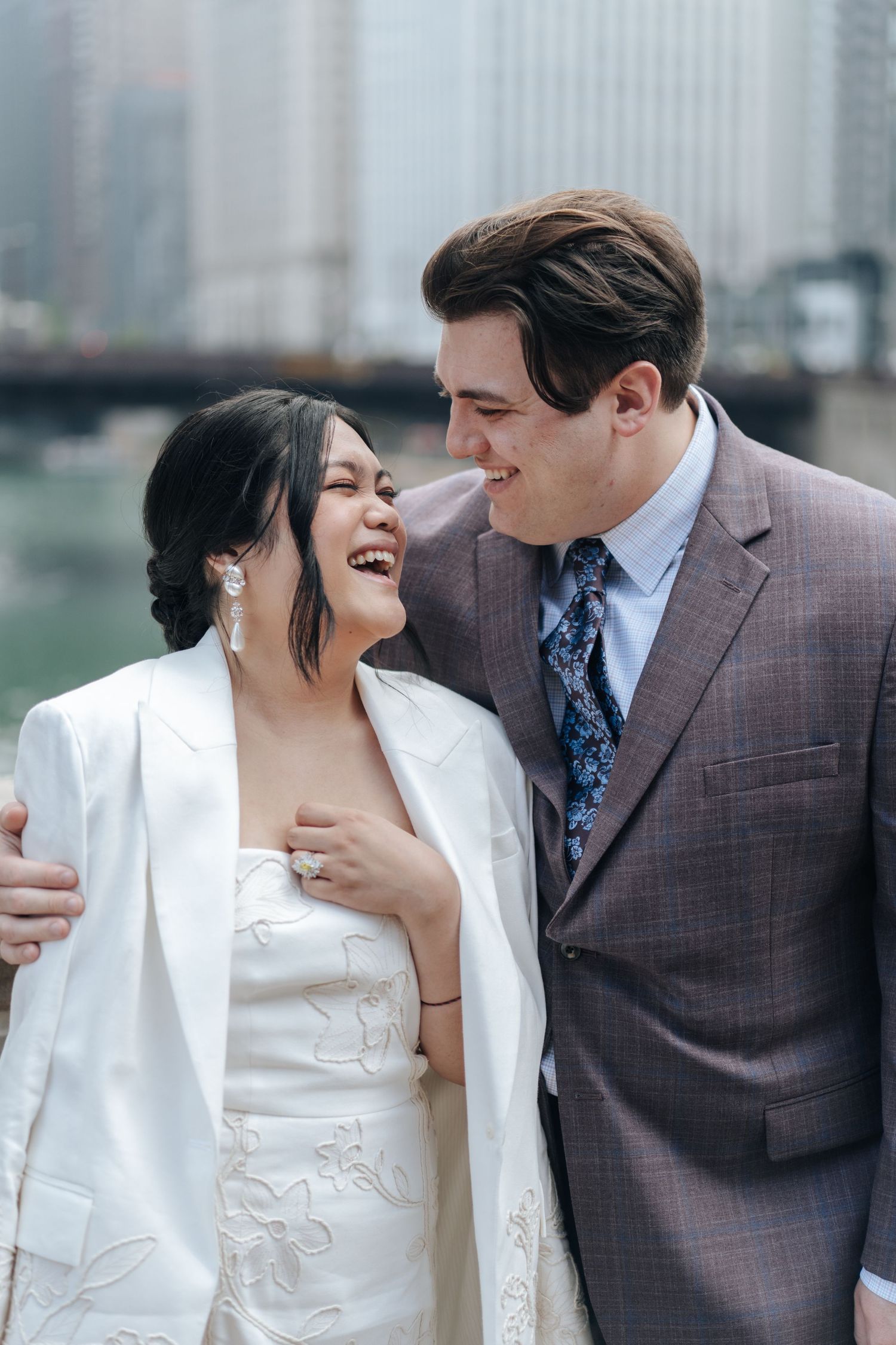 A laughing bride in a white dress and blazer with a groom in a purple suit share a joyful moment with a city skyline behind them.
