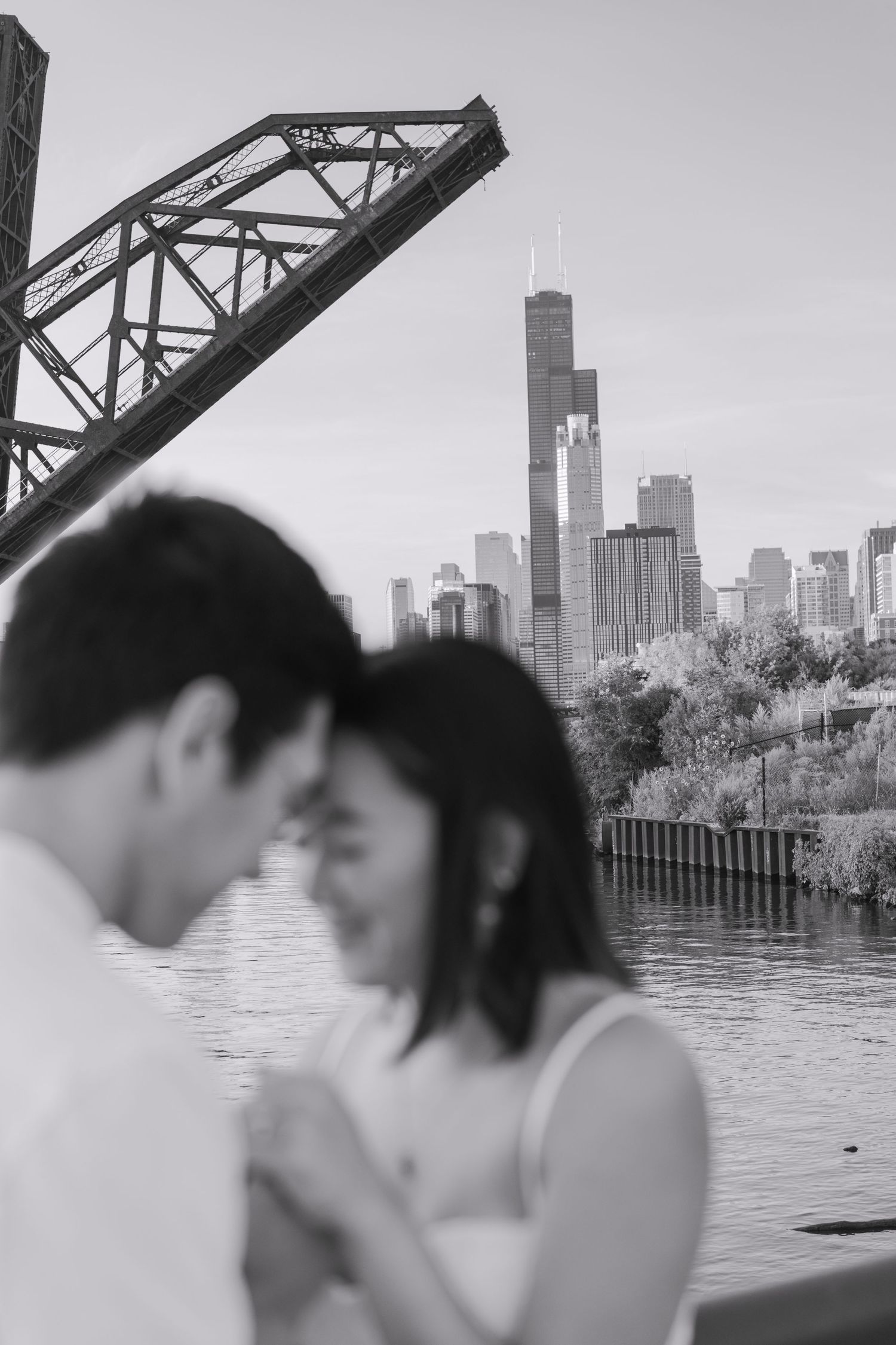 Black and white photo of a couple embracing near the Chicago River with Willis Tower and a raised bridge in the background.