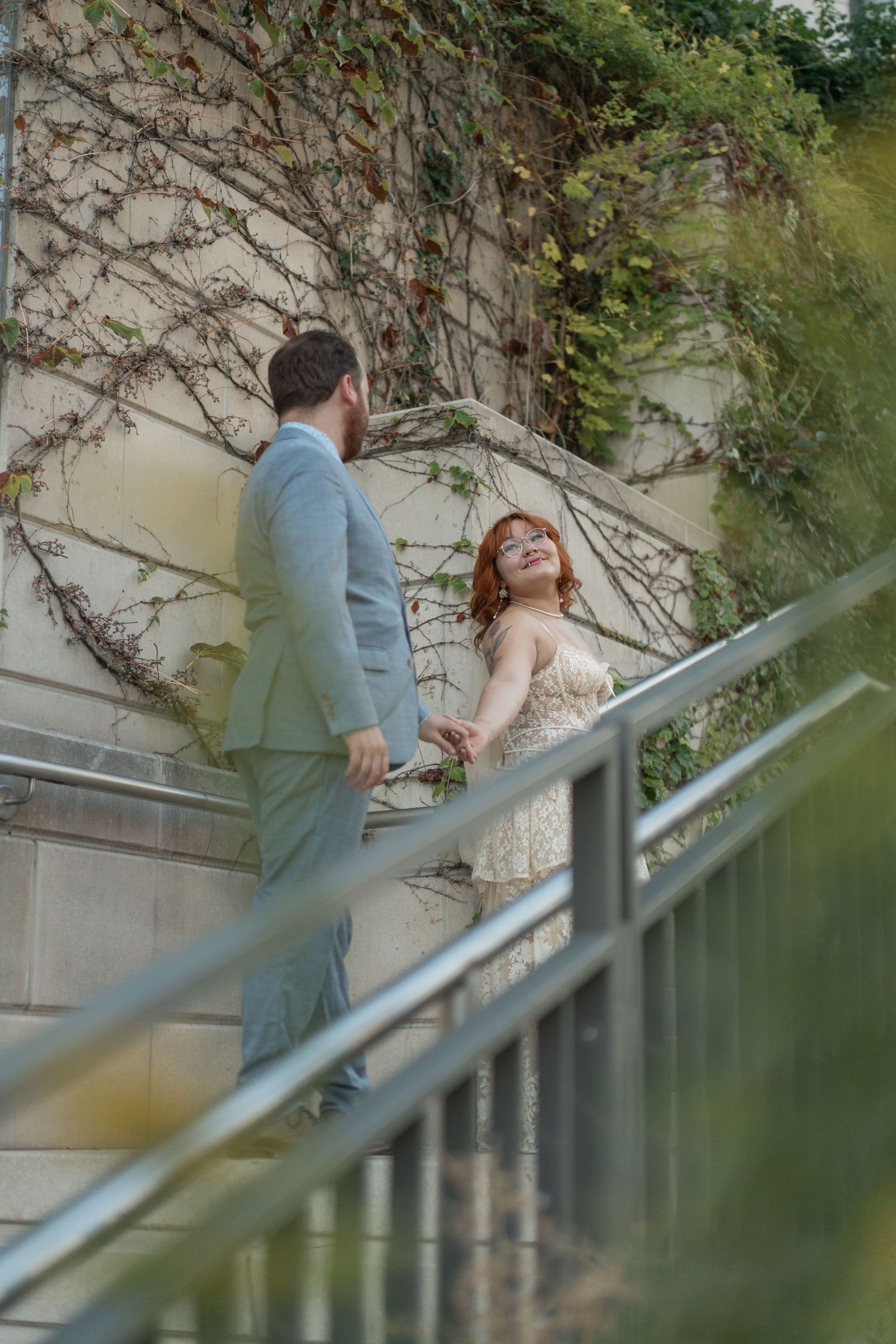 Bride and groom holding hands on outdoor stairs with ivy-covered wall, bride in lace gown smiling up at groom in grey suit.