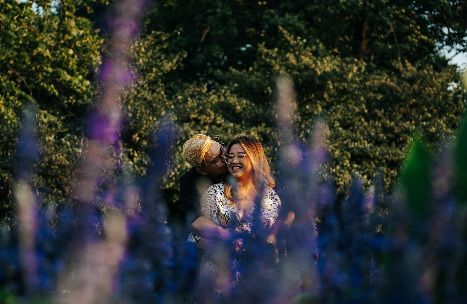 Couple sharing a romantic moment surrounded by vibrant purple wildflowers with lush green trees in the background.