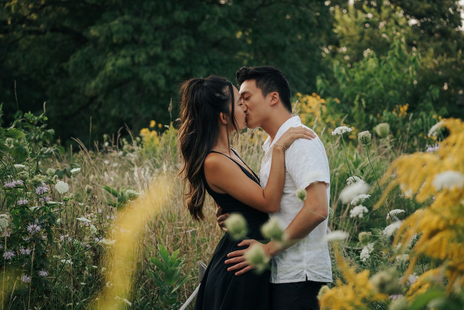A couple shares a romantic kiss surrounded by wildflowers and lush greenery in a scenic outdoor field.