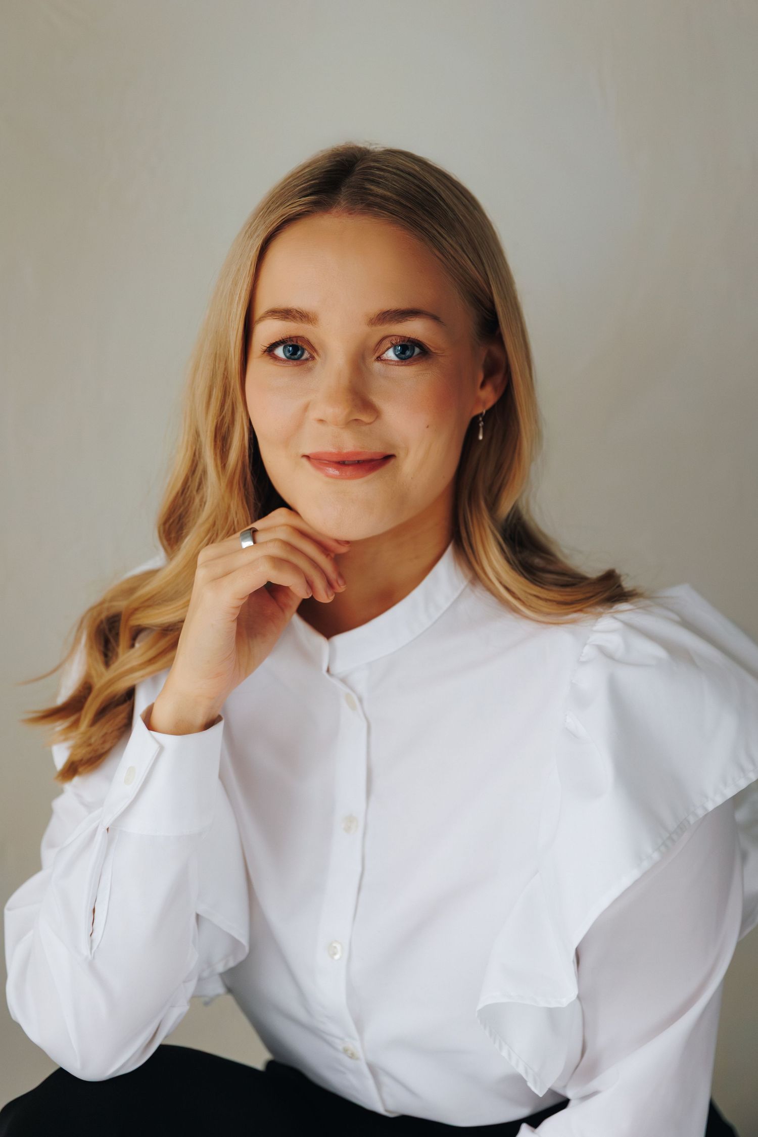 Portrait of a person with blonde wavy hair wearing a white button-up blouse with ruffled shoulders against a neutral background.
