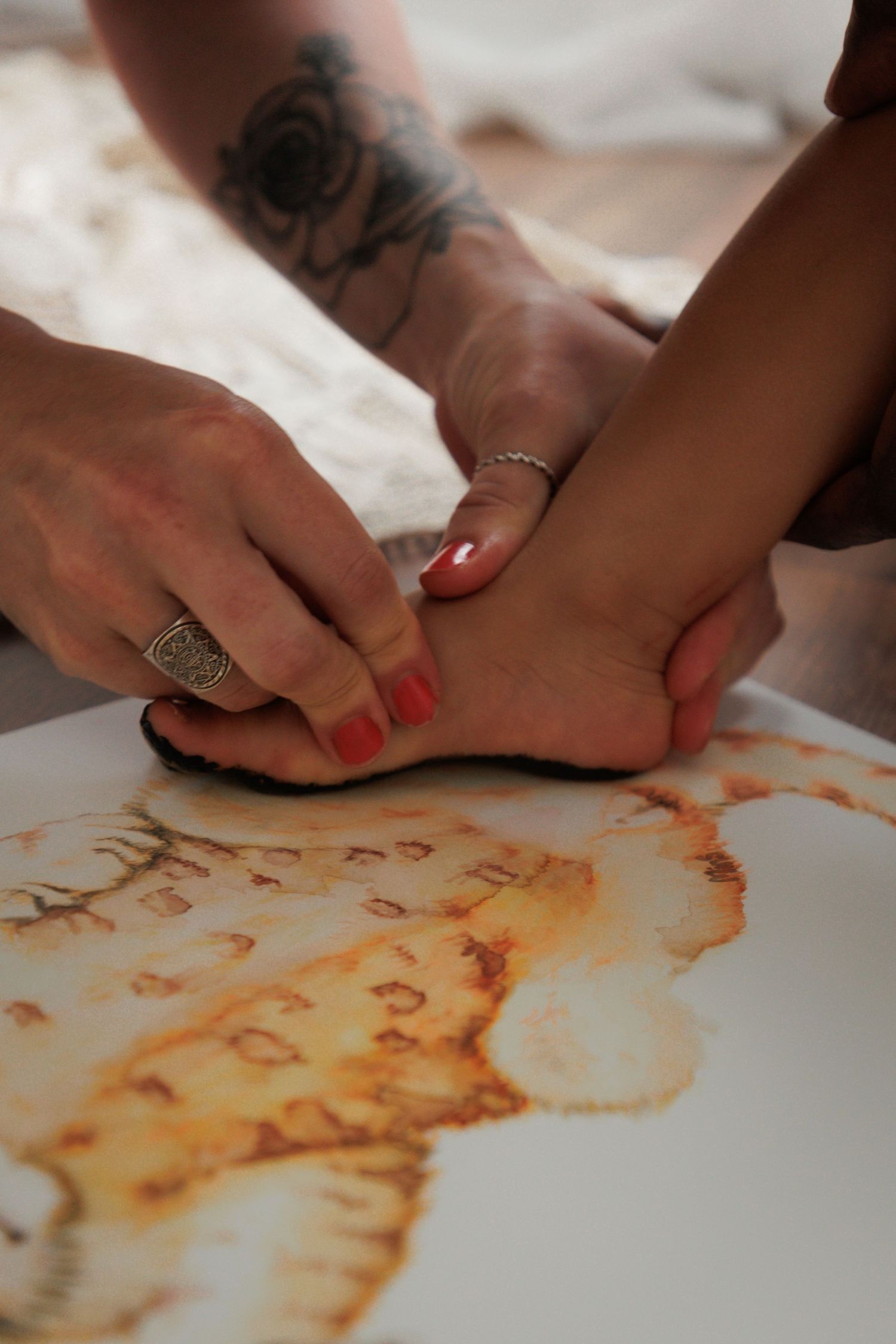Hands with rings and tattoos pressing a foot onto a surface with henna or paint print design.