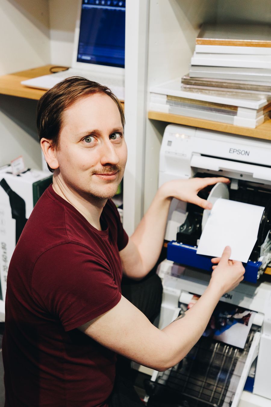 Man in red shirt smiling while operating an Epson printer in a print shop with stacked paper on shelves.