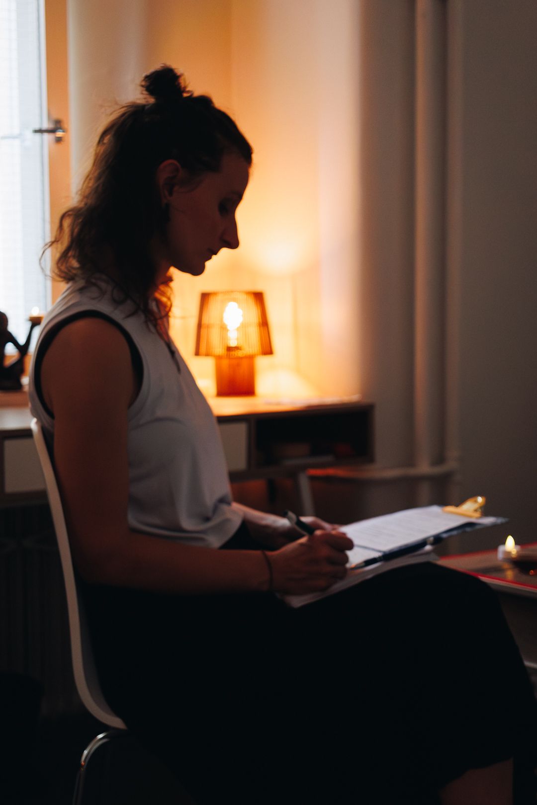 Woman sitting in dimly lit room writing on clipboard with warm lamp glowing in background.