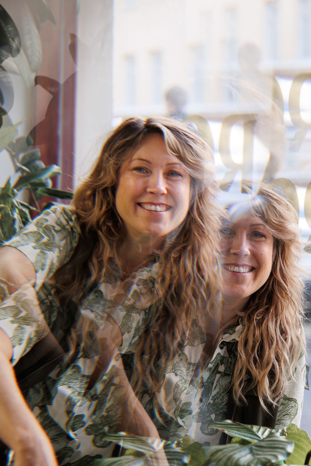 Two smiling women with long wavy hair pose together near green plants behind a window.