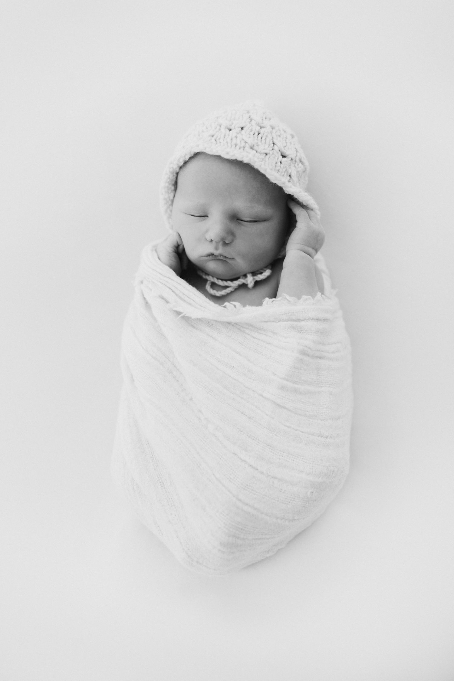 Black and white photo of a sleeping newborn baby swaddled in white wrap wearing a knit bonnet and pearl necklace.