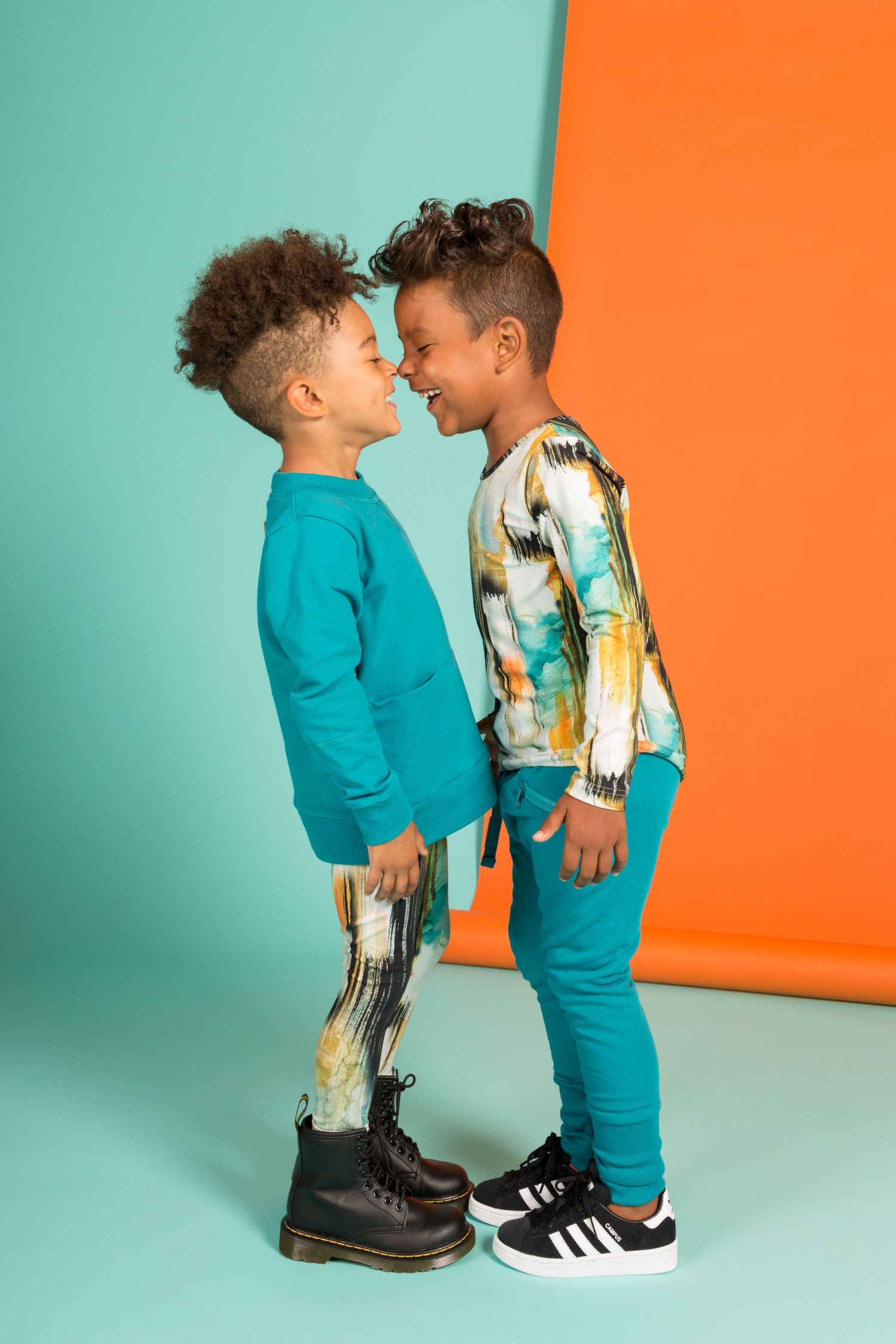 Two stylish kids in teal outfits playfully touch noses against a colorful orange and mint background.