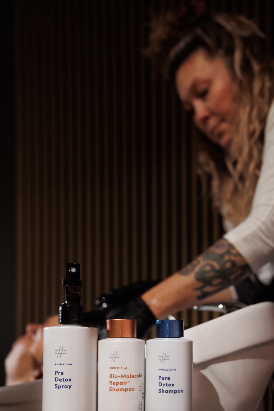 Tattooed hairstylist works behind a row of professional hair care products including Pre Detox Spray and Bio-Molecule Repair Shampoo.
