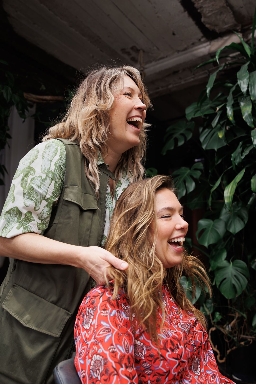 Two laughing women with wavy blonde hair enjoying a joyful moment outdoors near lush green tropical plants.