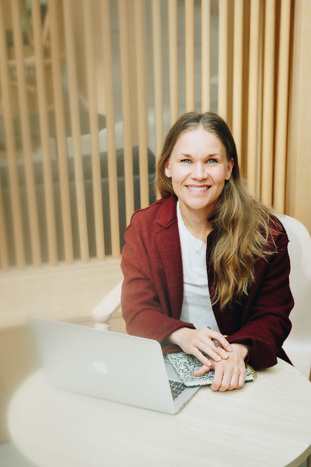 Happy woman in red blazer smiling at camera while working on laptop in bright office.