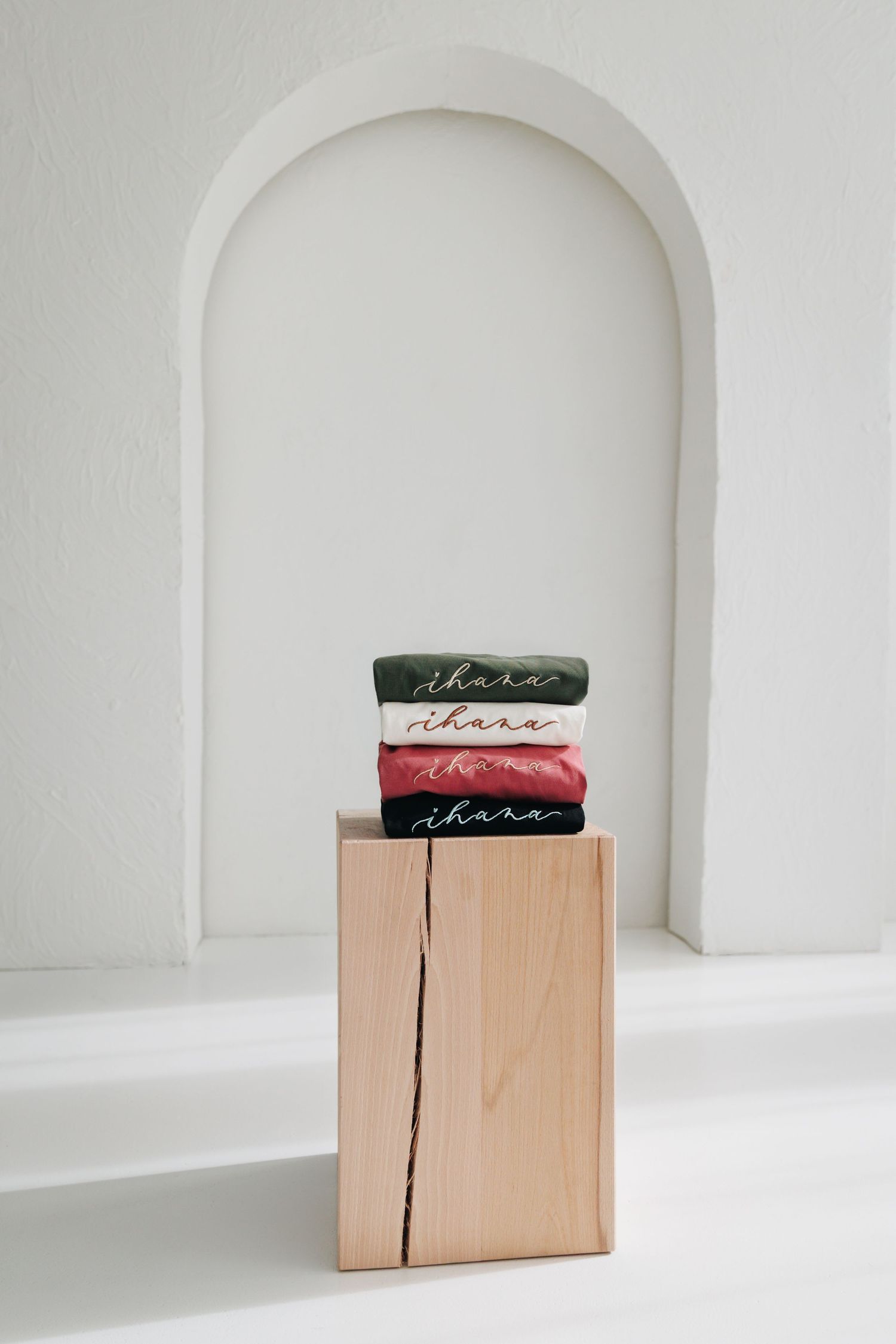 Stack of folded ihara branded shirts in green, red, and neutral tones displayed on a wooden block pedestal.