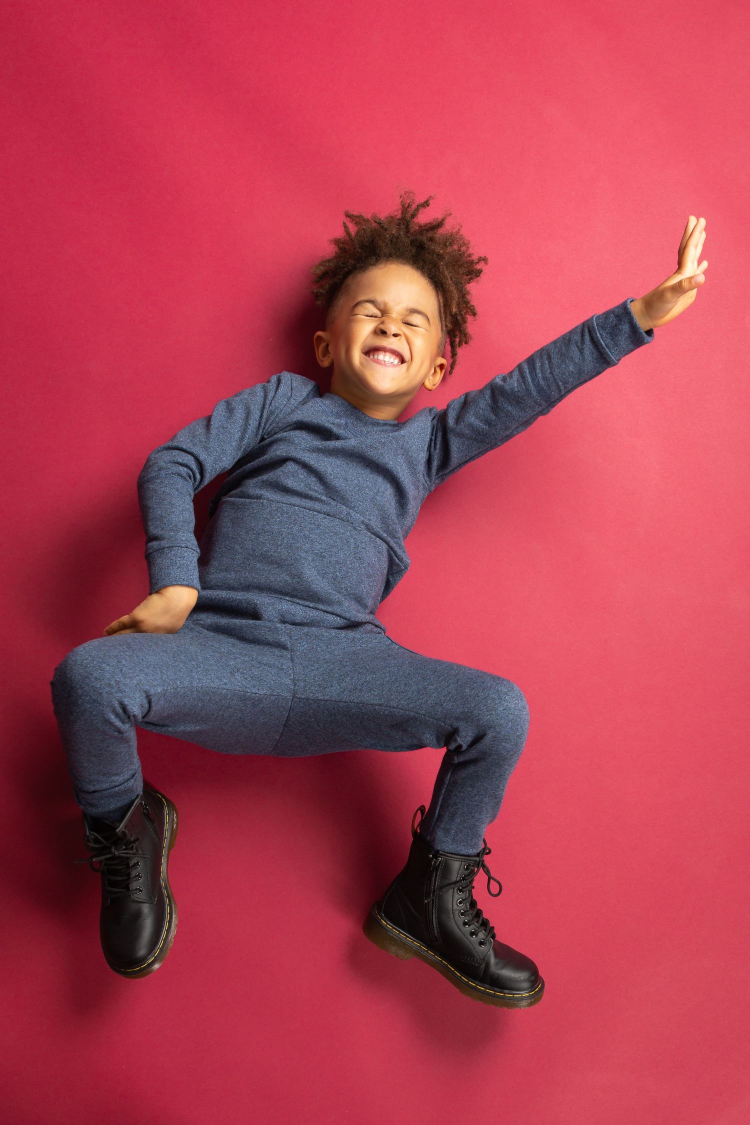 Joyful Black child in blue tracksuit and boots jumping against a red background with arms outstretched.