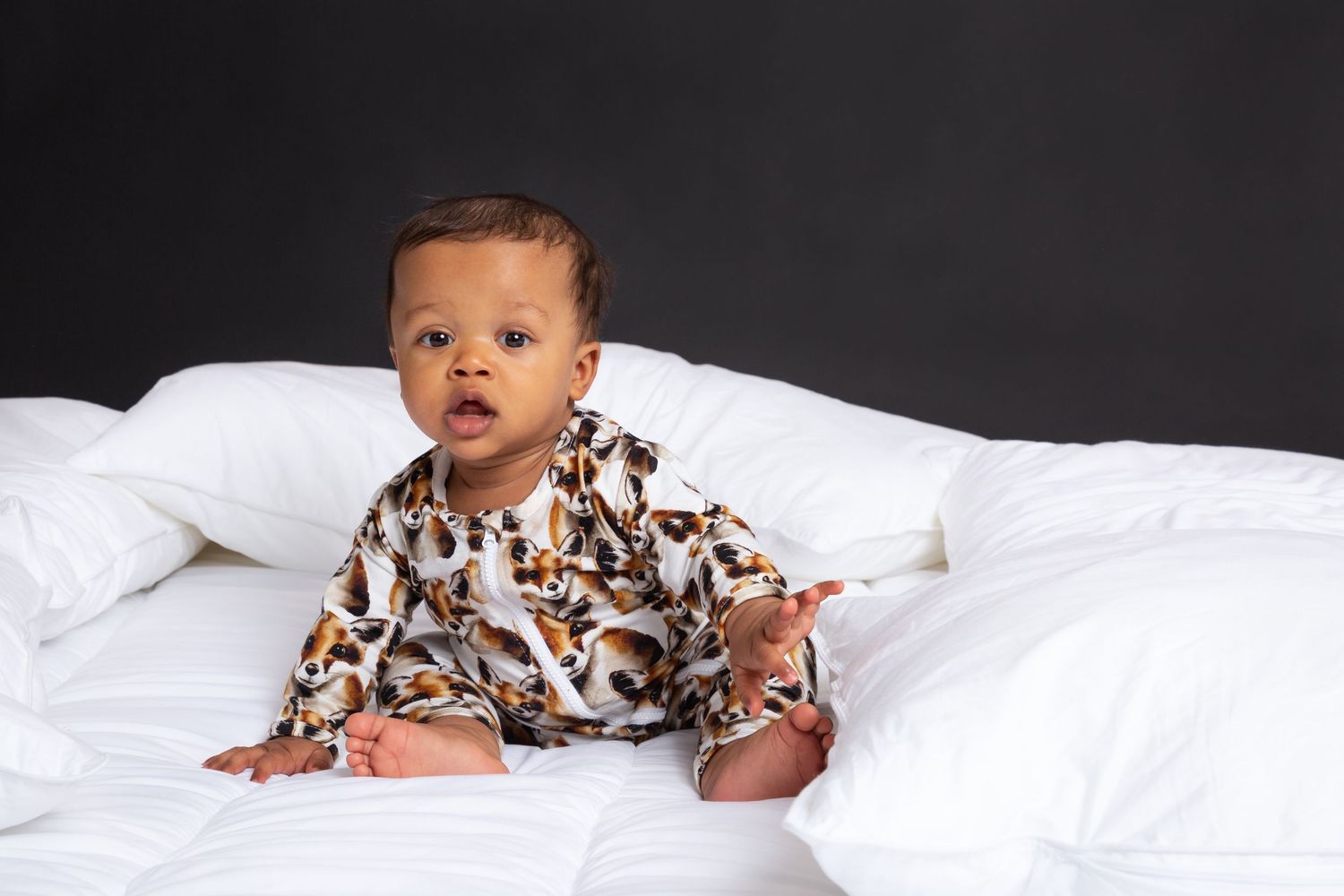 Baby in leopard print pajamas sitting on white bedding against dark background.