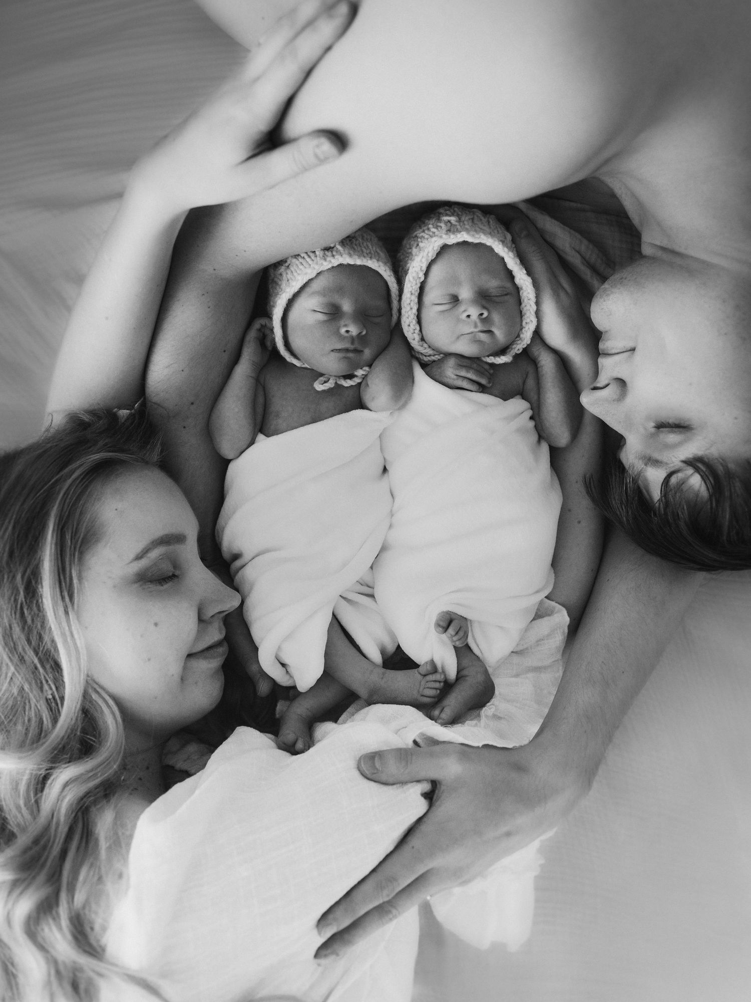 Black and white photo of two newborn twins in bonnets lying between their parents on a bed.