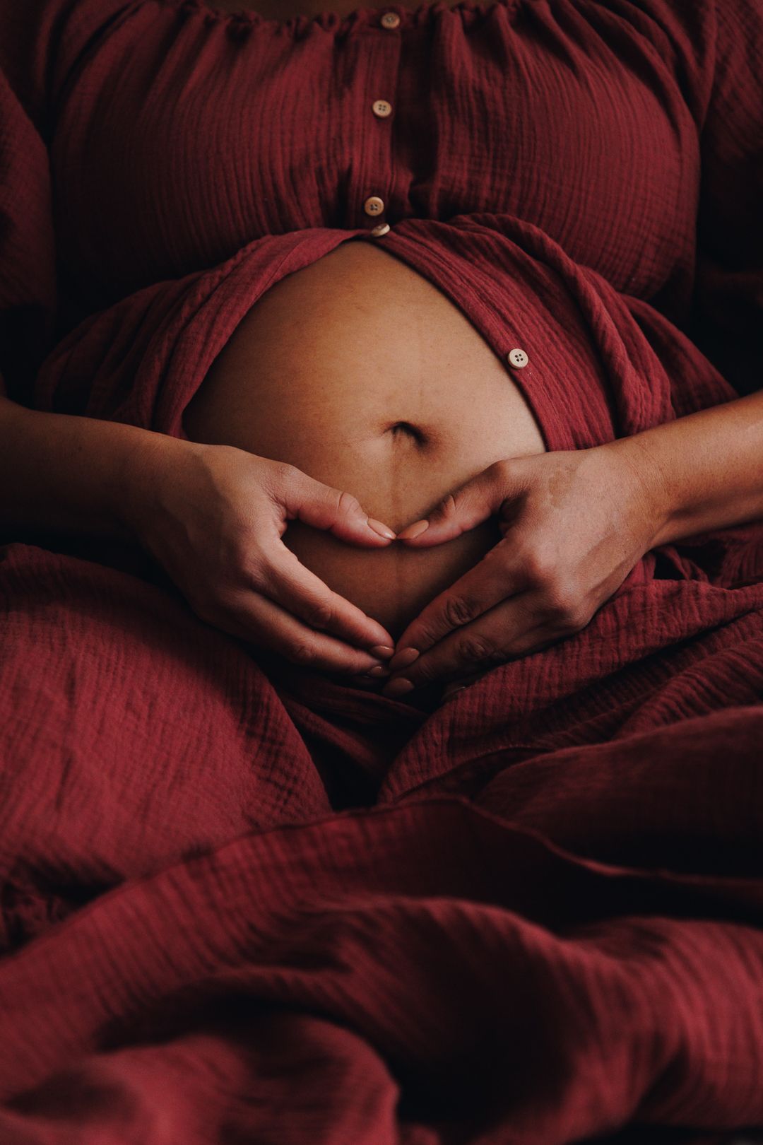 Pregnant woman's belly in burgundy dress with hands gently cradling her stomach in intimate maternity photo.