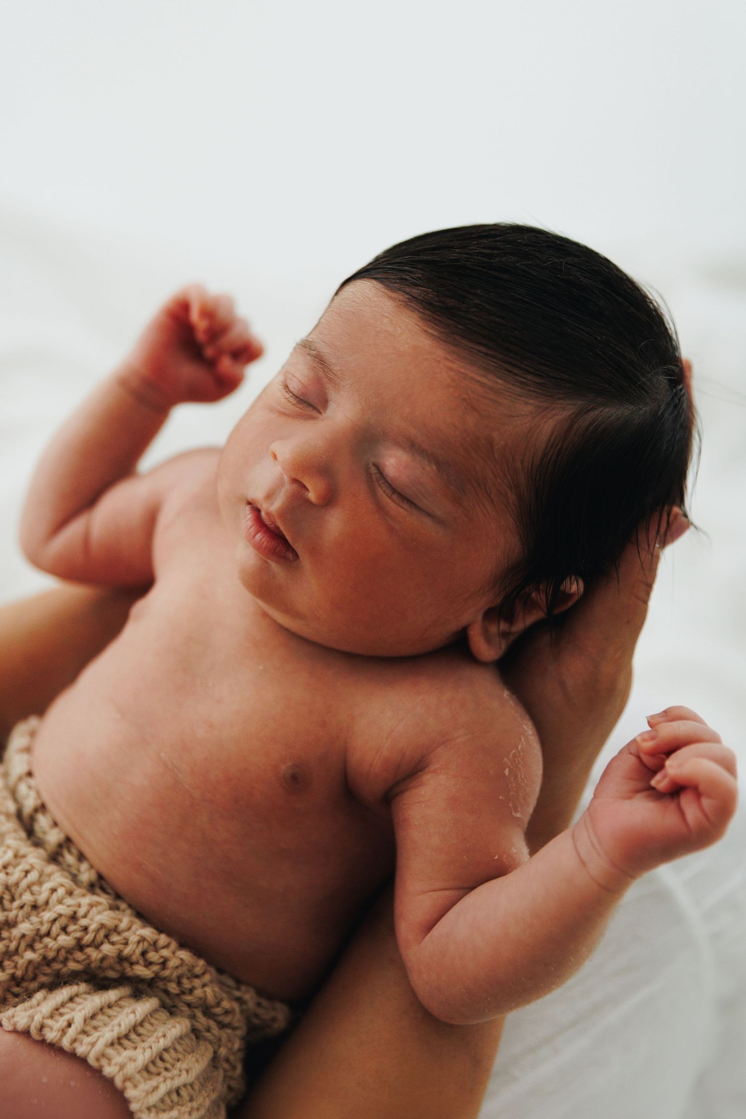 Peaceful newborn baby with dark hair sleeping in knit diaper cover, arms raised, cradled in soft white background.