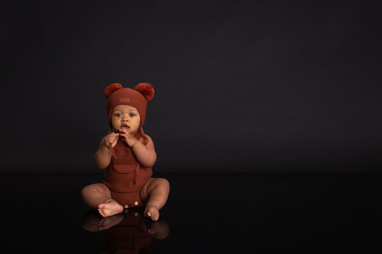 Baby in rust bear hat and onesie sits on dark reflective floor, hands clasped, studio portrait.