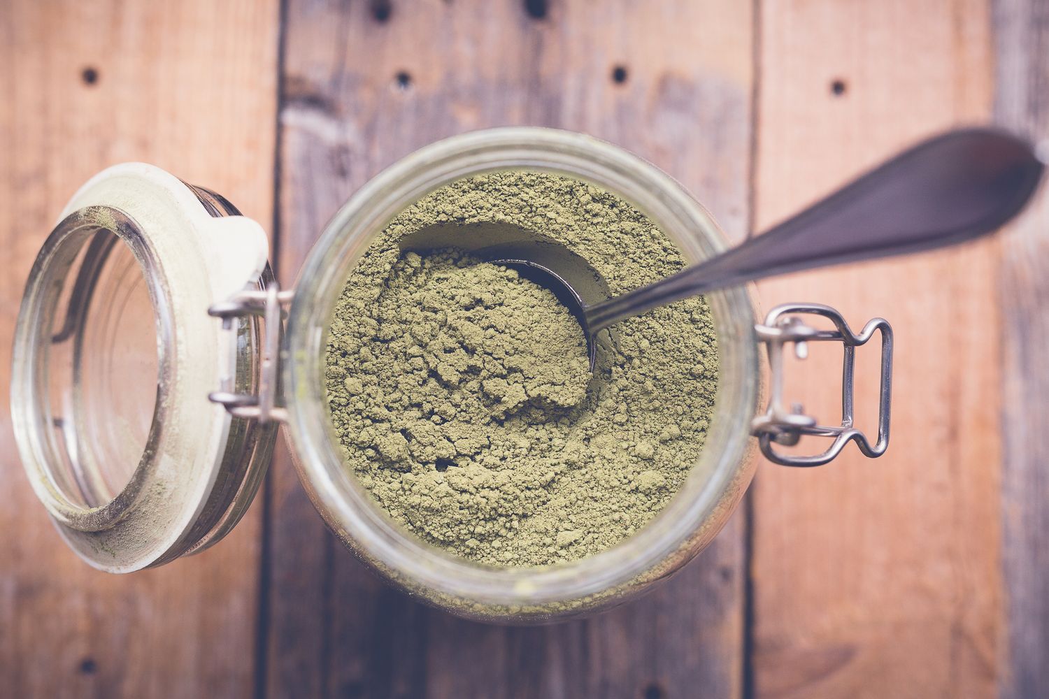Overhead view of green herbal powder in a glass jar with a metal spoon on a rustic wooden surface.