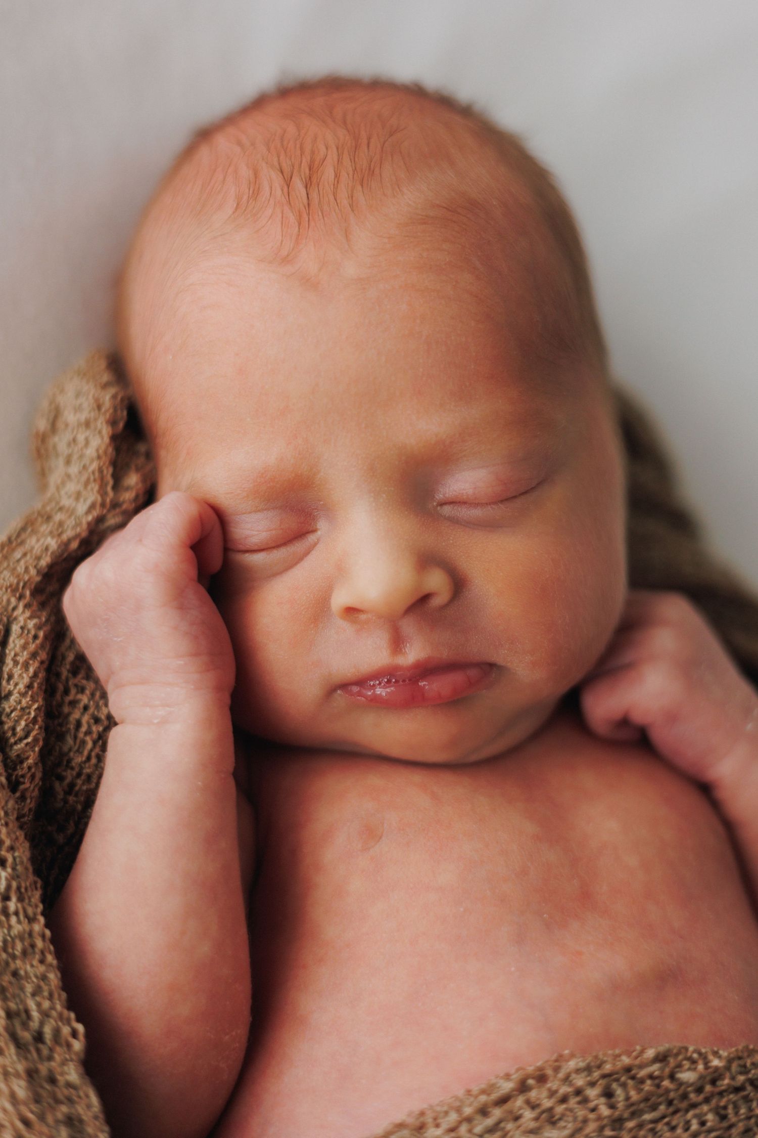 Peaceful newborn baby sleeping wrapped in a brown knit blanket with tiny hand resting against cheek.