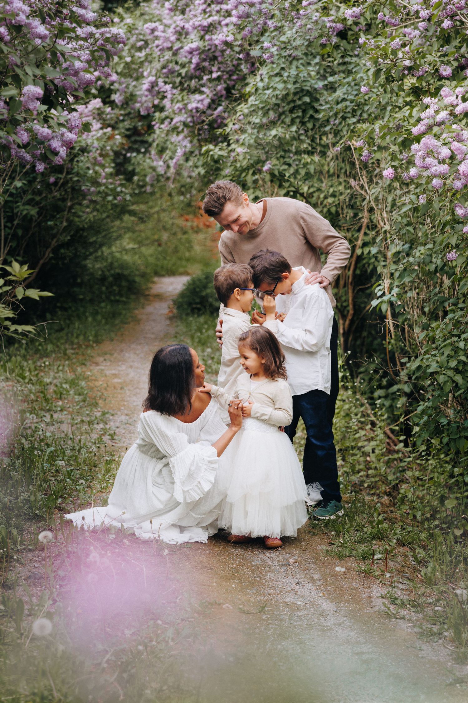 Family of four in white outfits posing among blooming lilac bushes on a garden path.