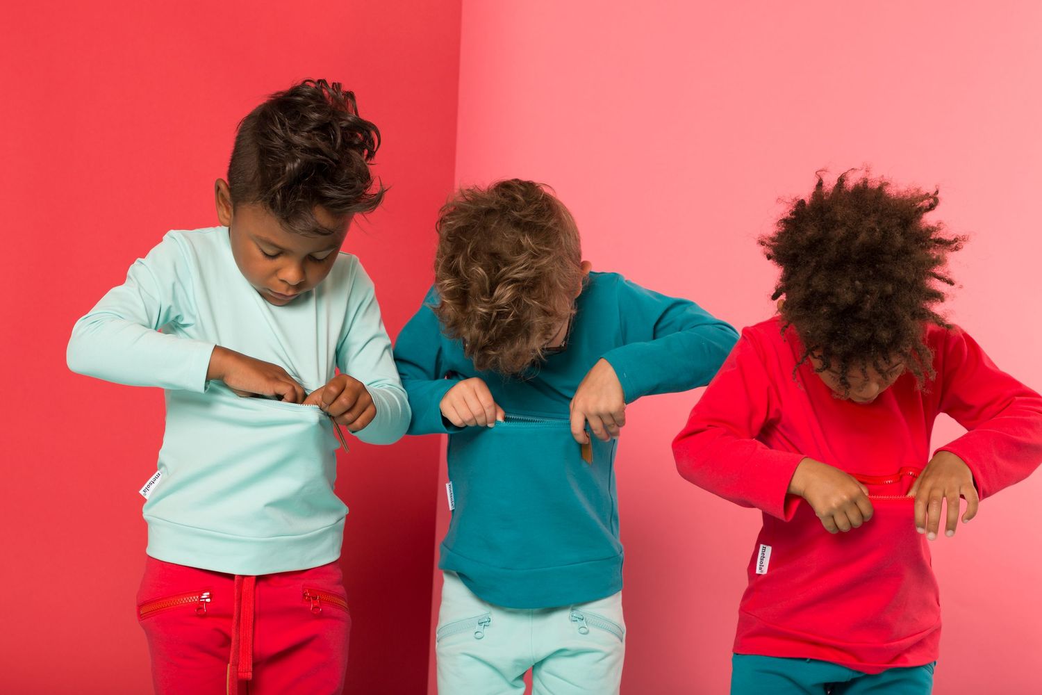 Three children in colorful sweatshirts dancing against a pink background, wearing red and teal outfits.