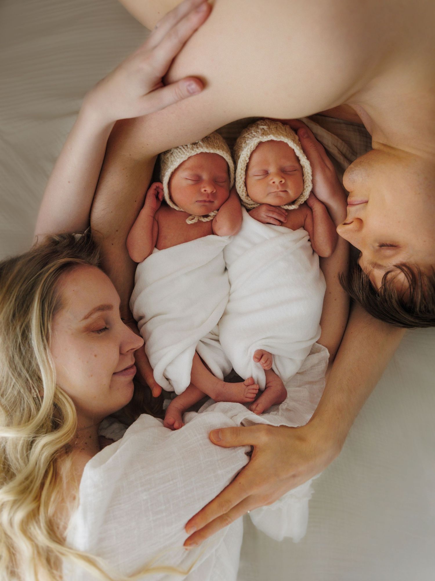Two parents lying down with newborn twins wrapped in white swaddles wearing knit bonnets between them.