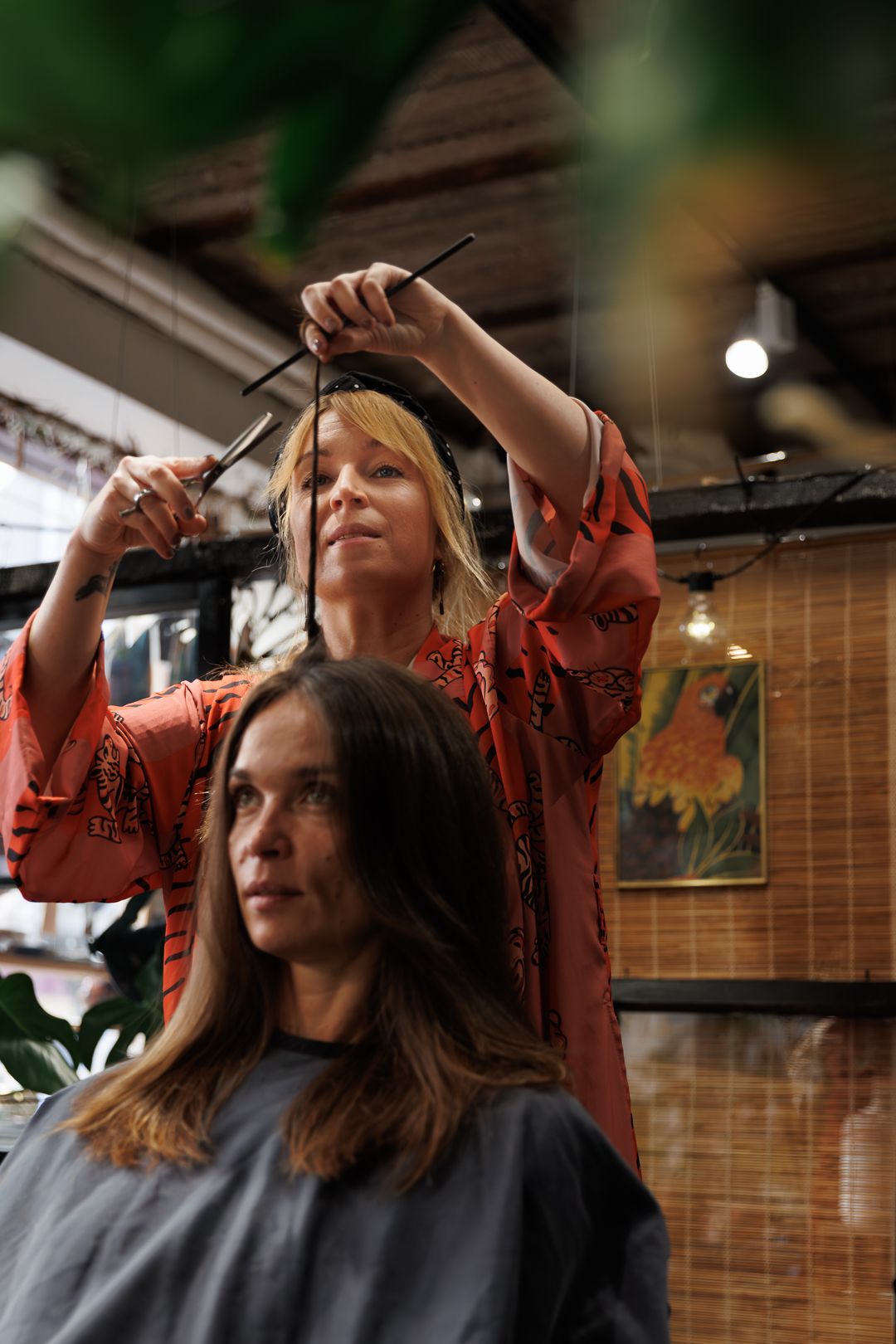 Blonde hairstylist in red kimono cutting brunette client's hair in a stylish salon.