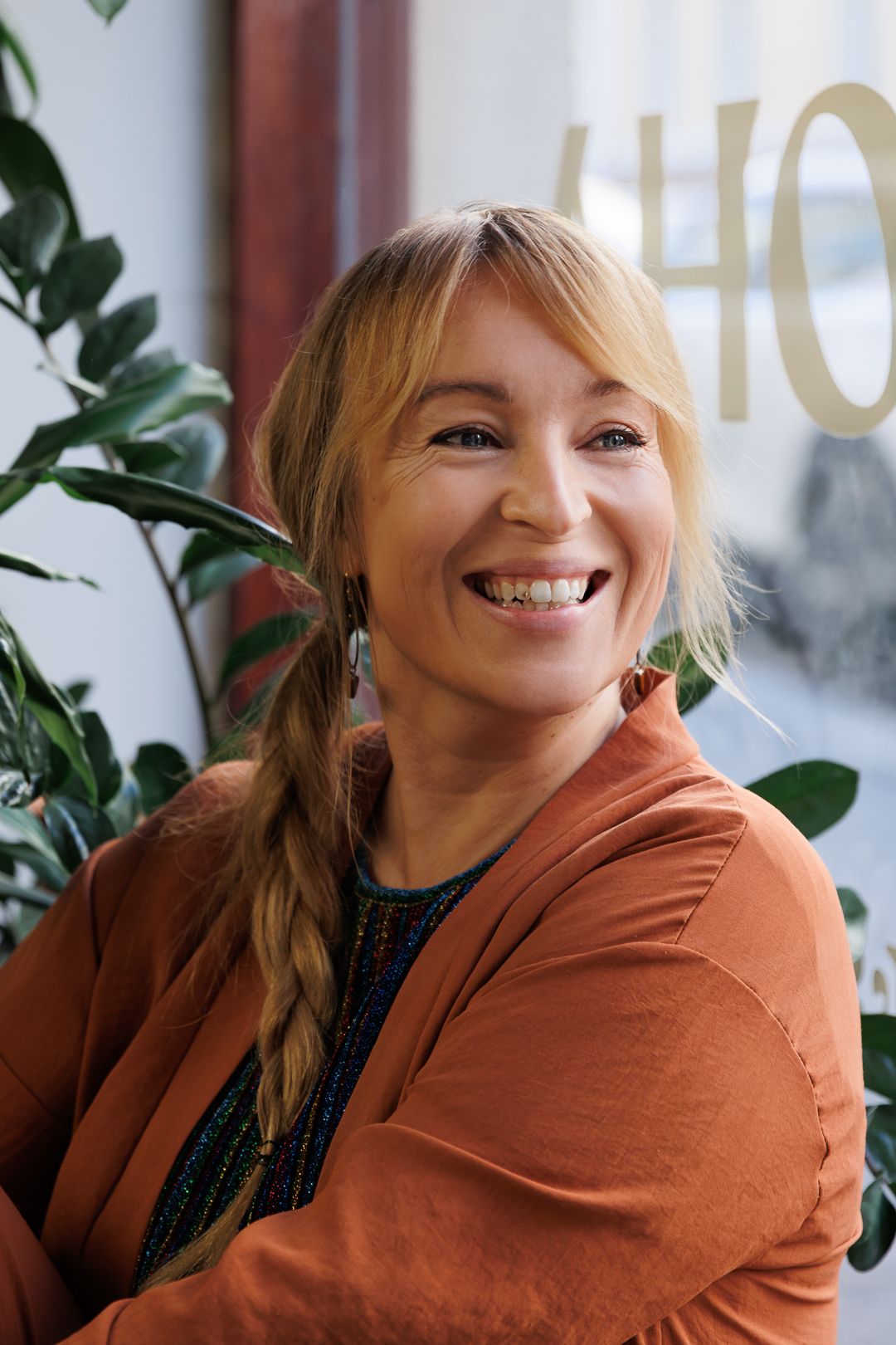 Cheerful blonde woman with braid wearing orange blazer smiles in bright salon with green plants.