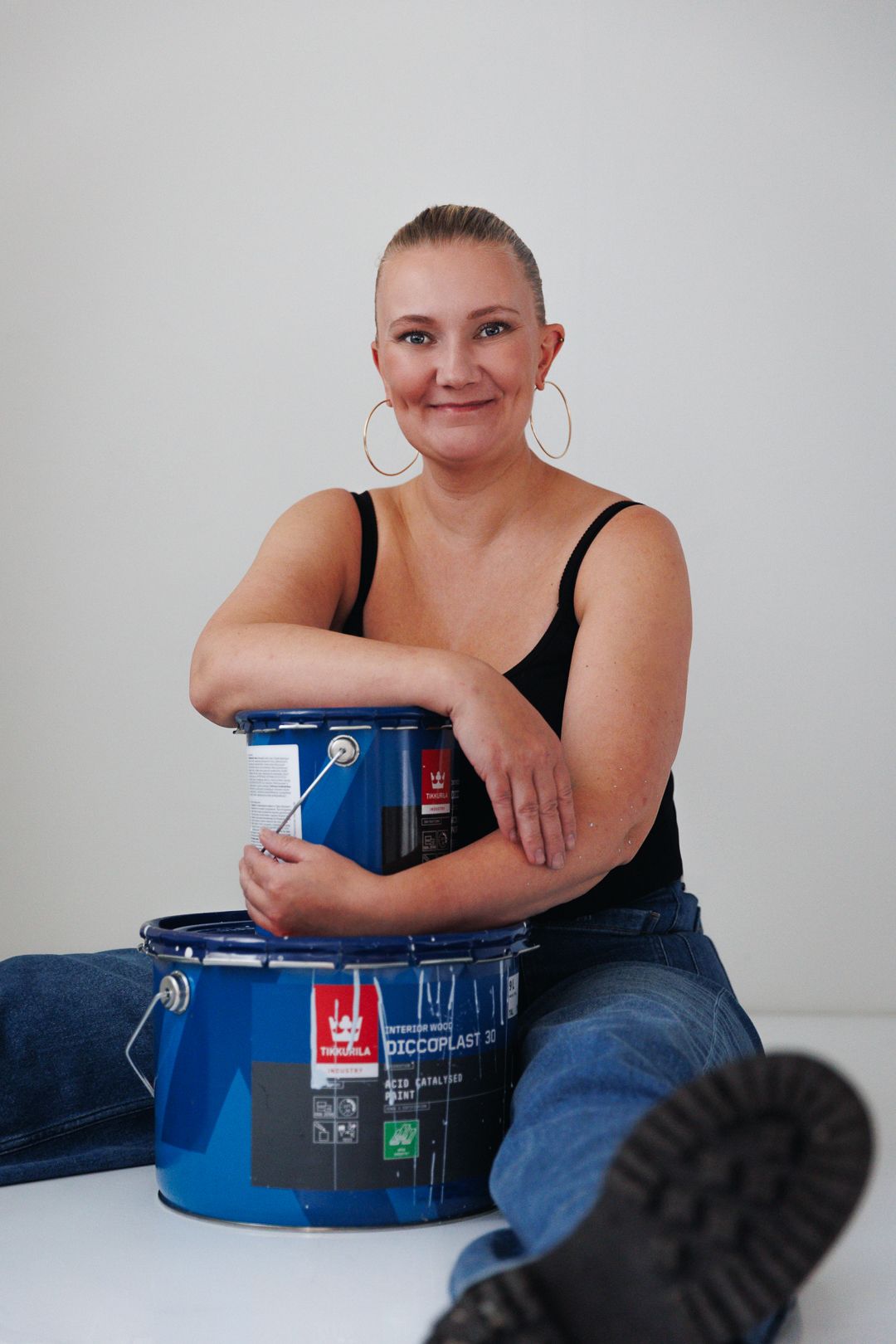 Woman in black tank top and jeans sitting with a blue Tikkurila paint can, smiling at camera.