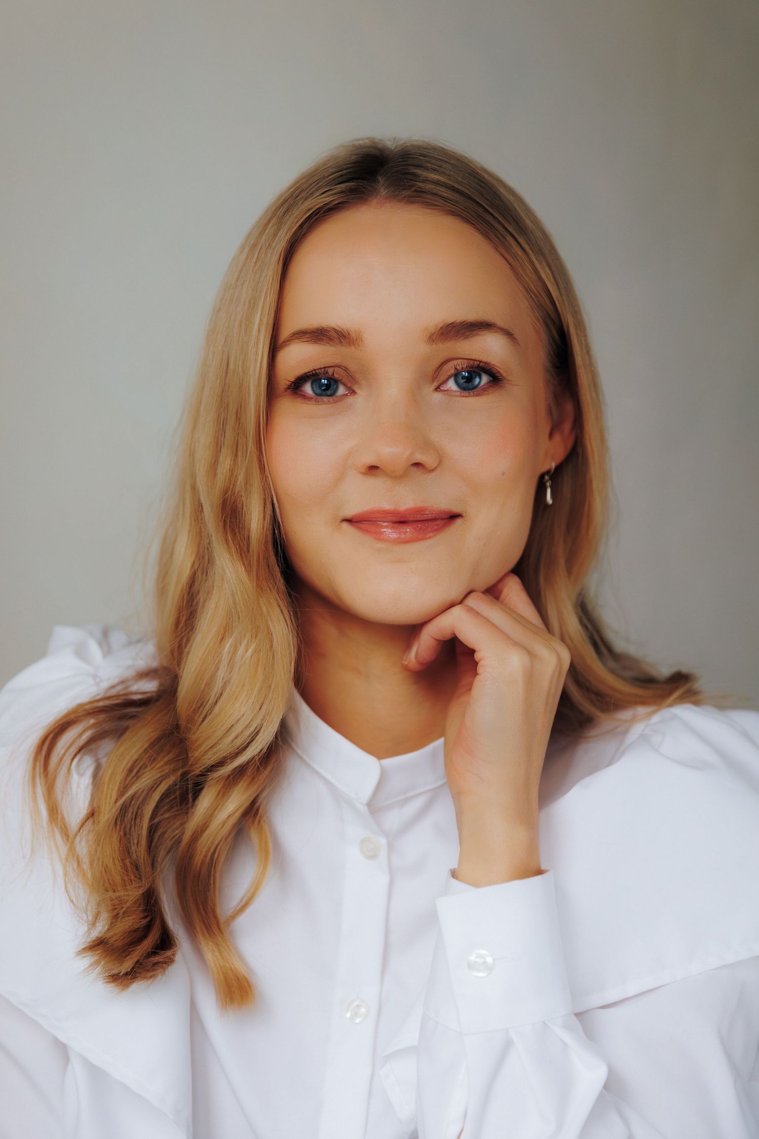 Young blonde woman in white ruffled shirt smiling softly, resting chin on hand against light gray background.