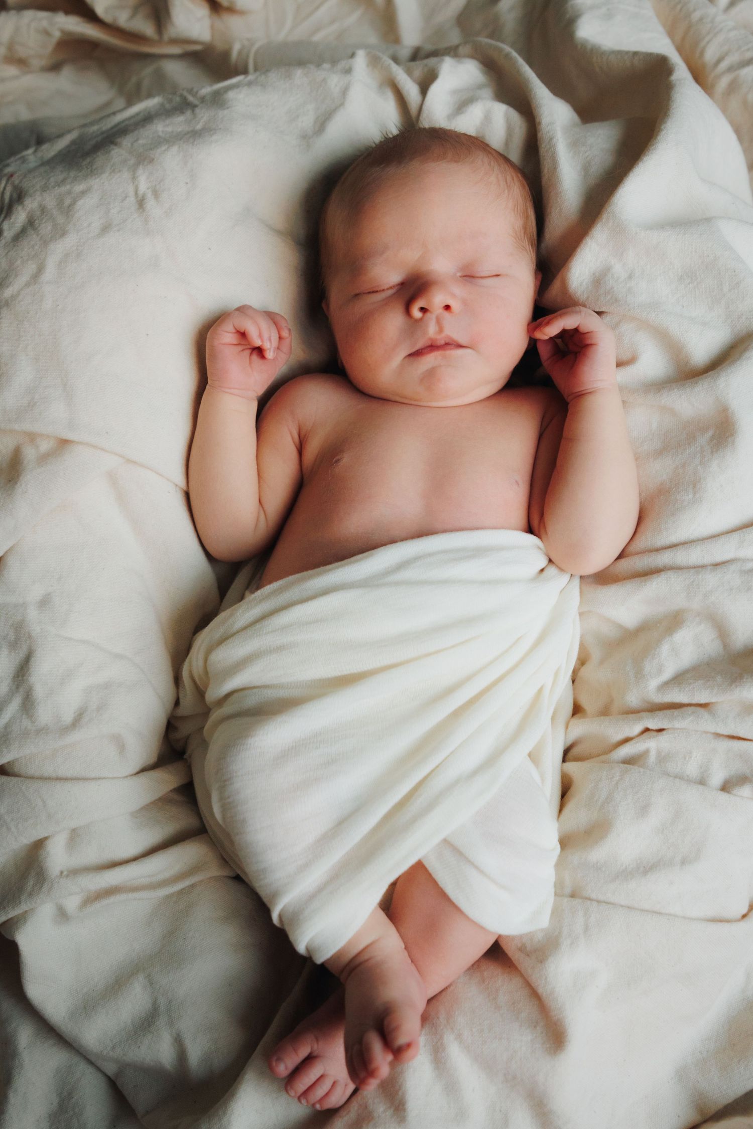 Newborn baby wrapped in soft white fabric sleeping peacefully on white bedding sheets.