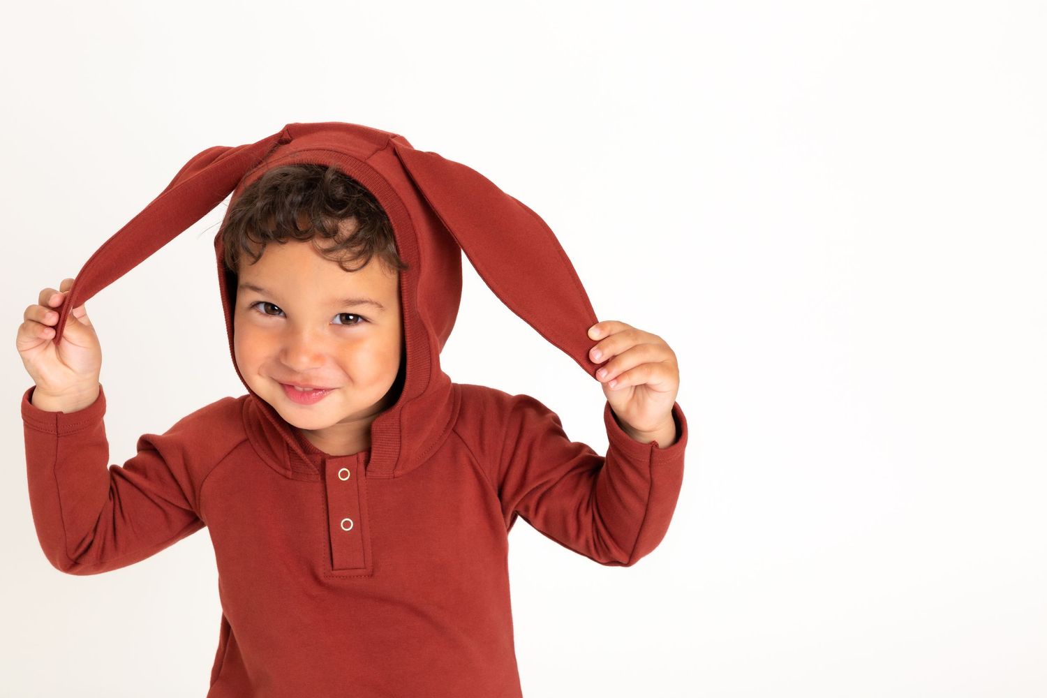 Young child wearing red hooded top playfully pulls on the hood's ear-like flaps while smiling against white background.