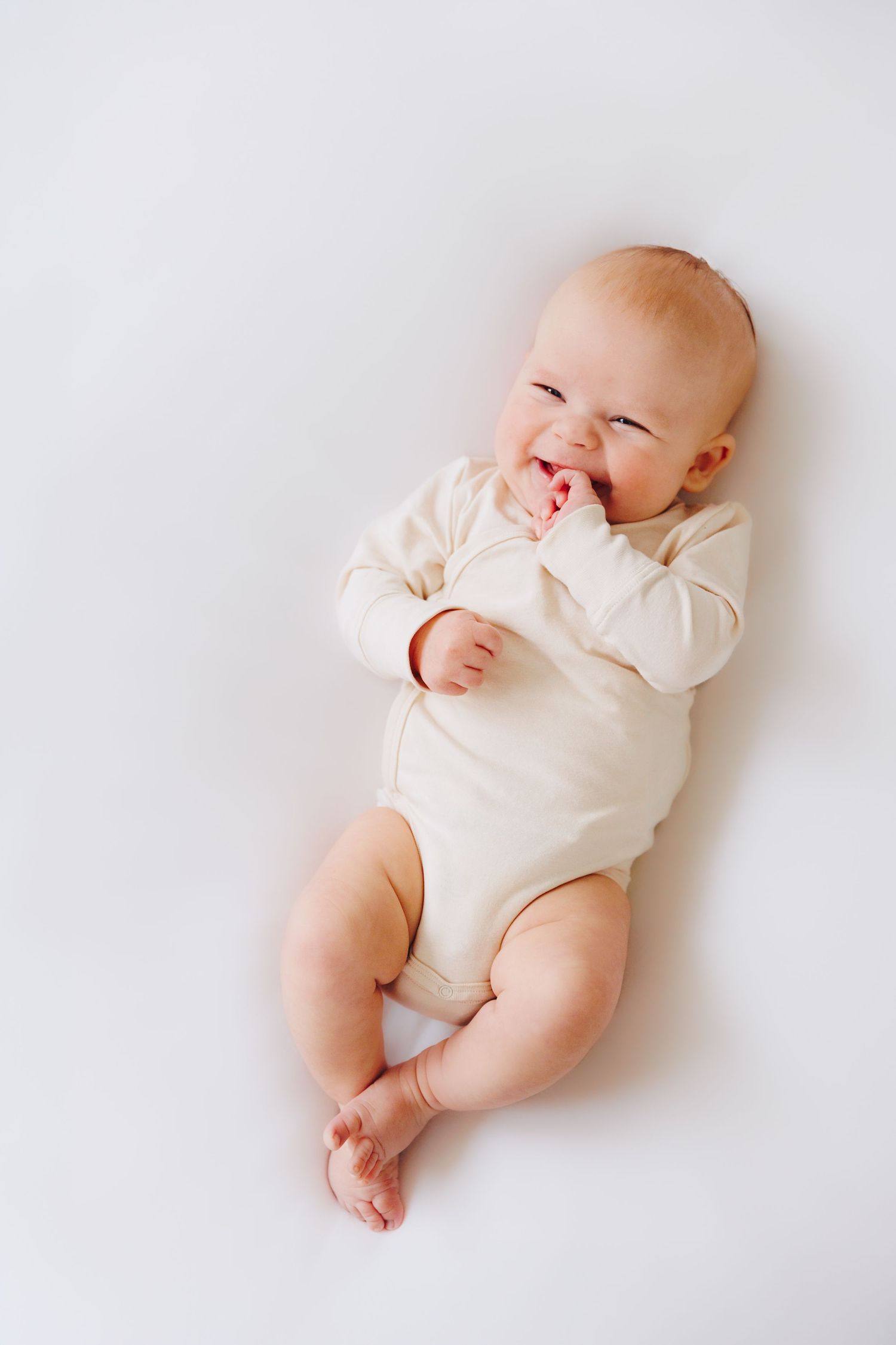 Laughing baby in cream bodysuit lying on white background.