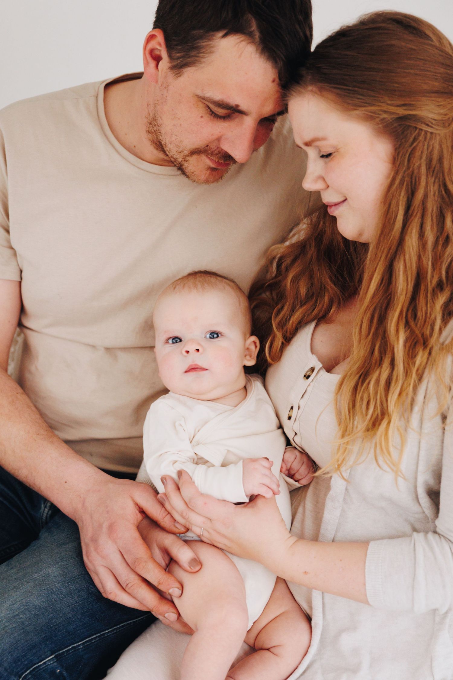 A young couple with red and brown hair lovingly gazes at their blue-eyed baby in white onesie.