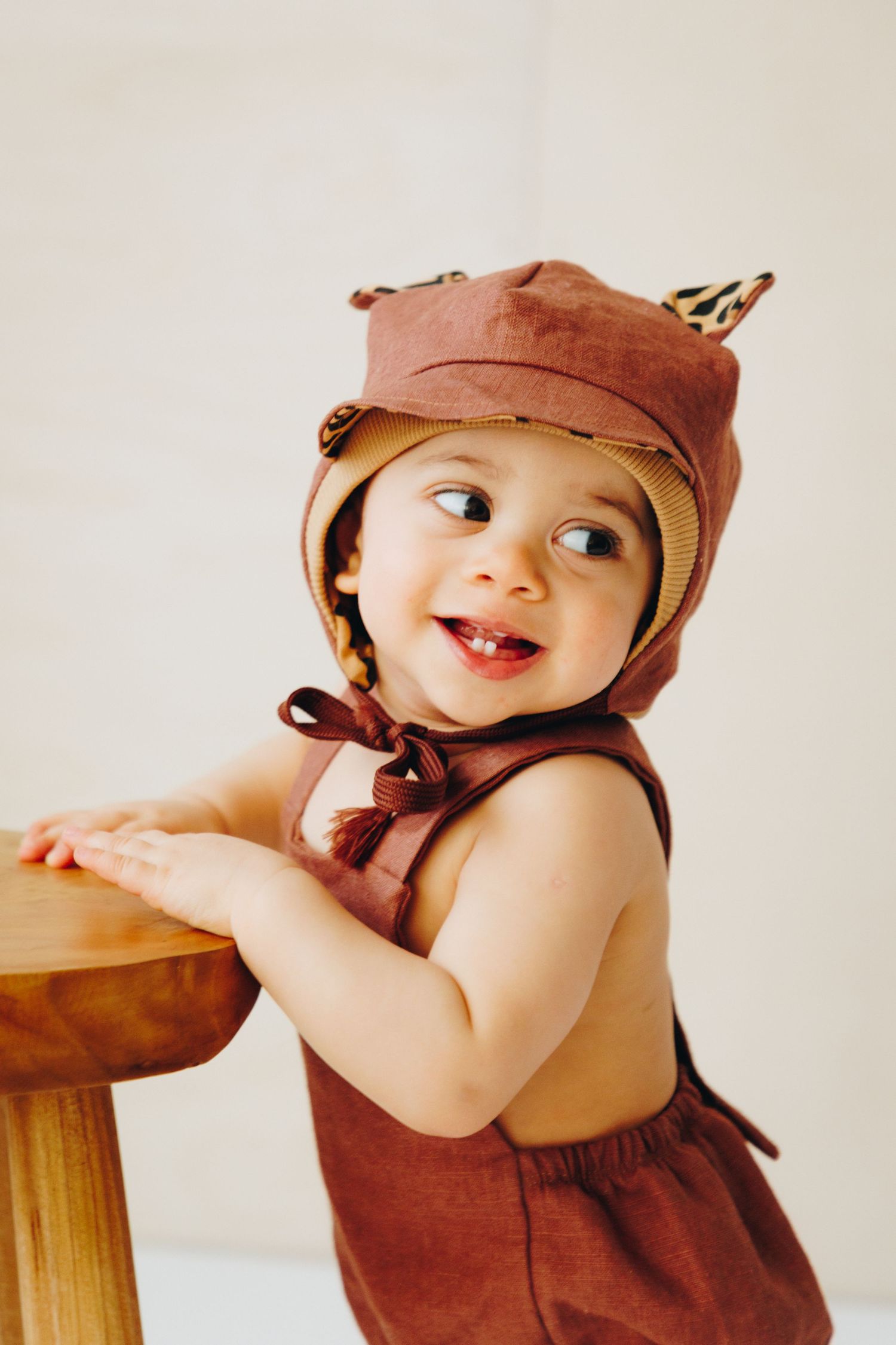 Adorable baby in brown romper and animal ear bonnet hat smiling while standing at a wooden stool.