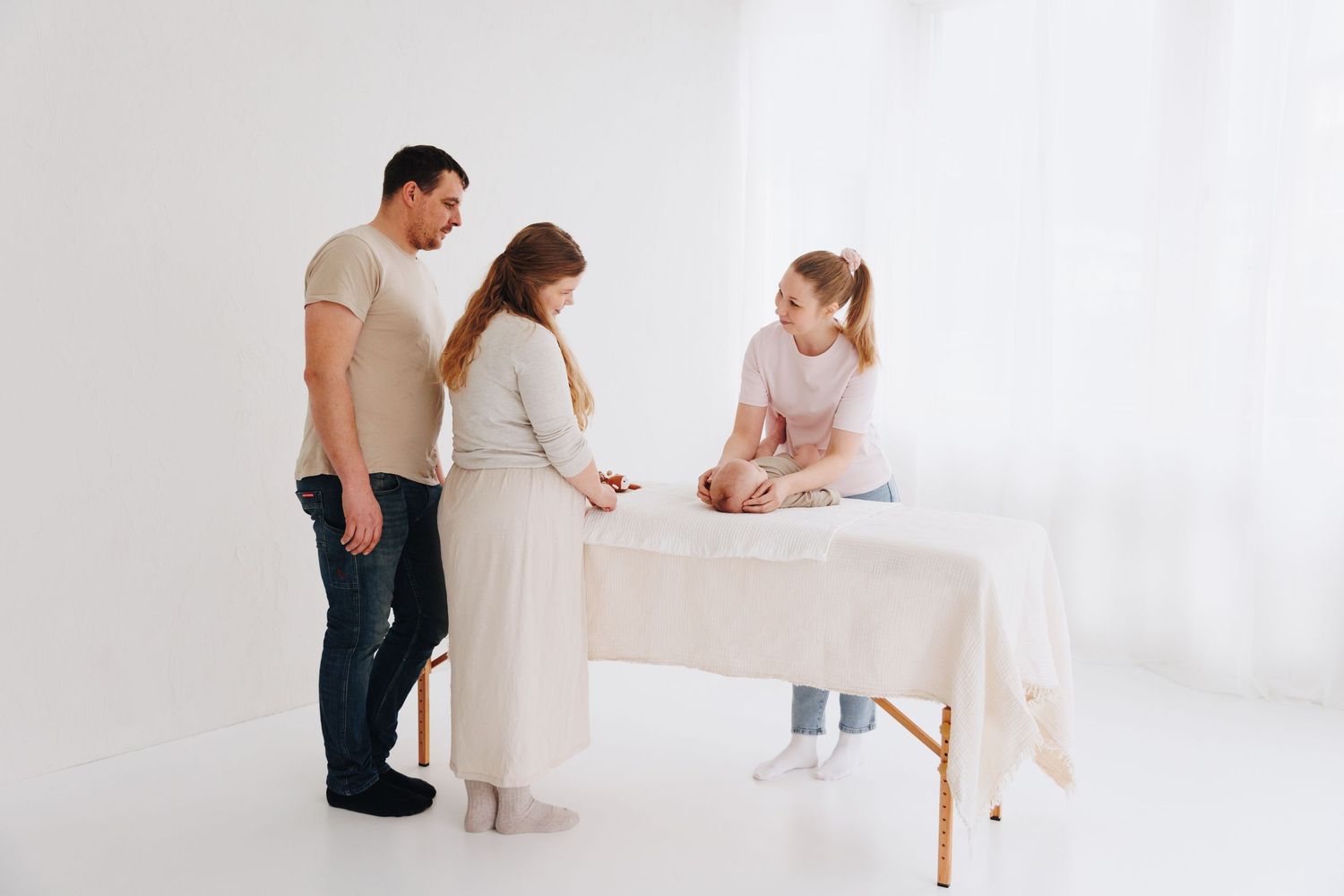 Parents and therapist examine newborn baby on massage table in bright white medical office setting.