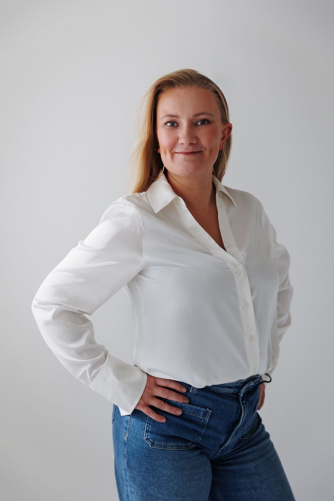 Woman in white button-up shirt and jeans posing confidently with hands on hips against light background.