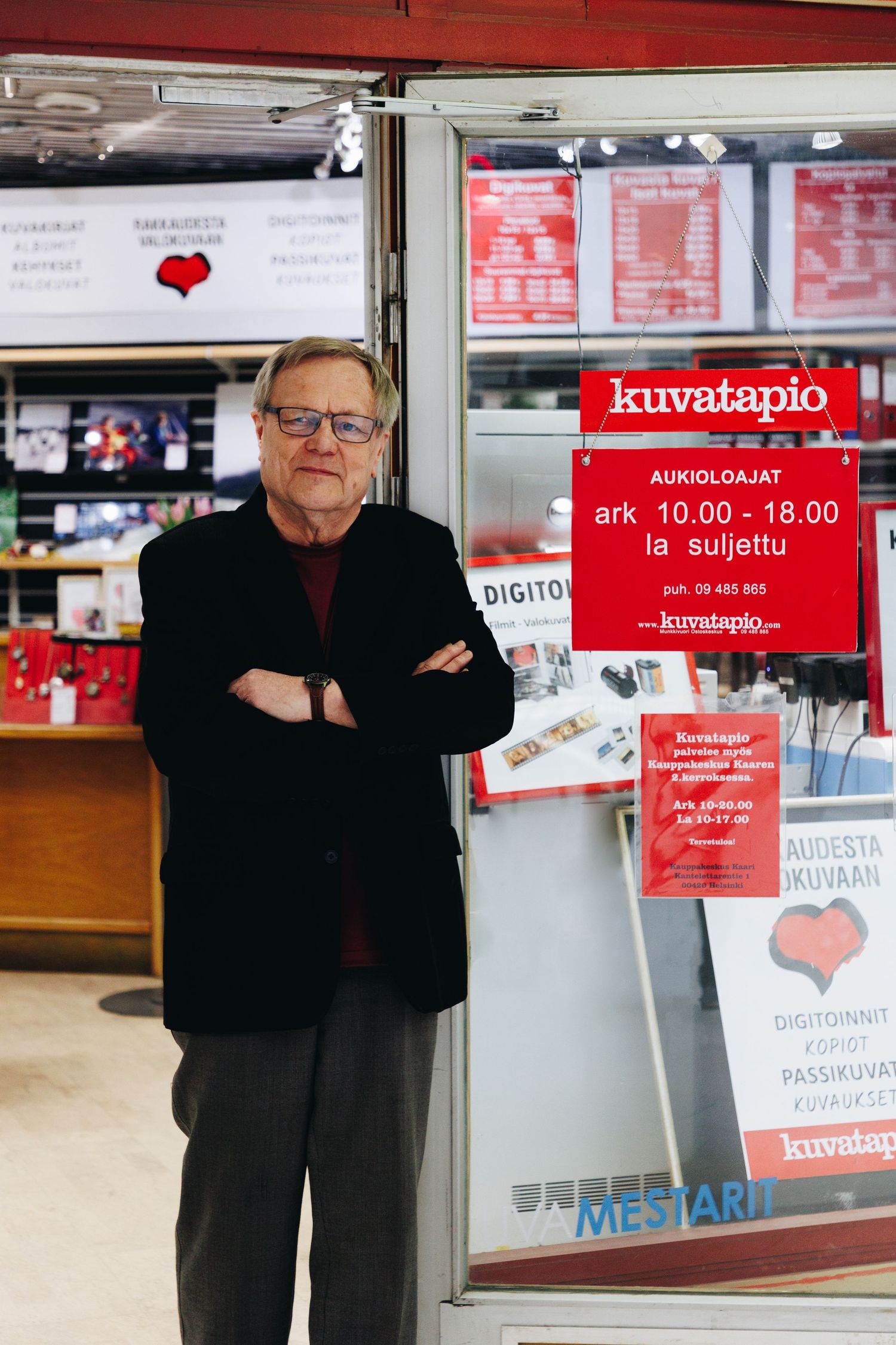 Elderly man with glasses stands arms crossed outside Kuvatapio photo shop in Finland displaying opening hours sign.