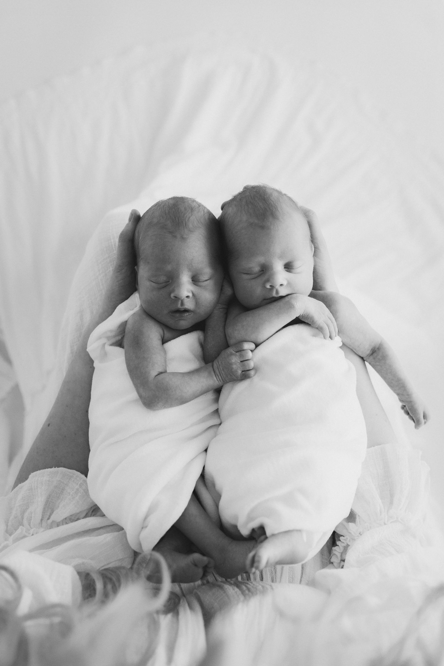 Black and white photo of two sleeping newborn twins swaddled in white blankets, gently nestled together.