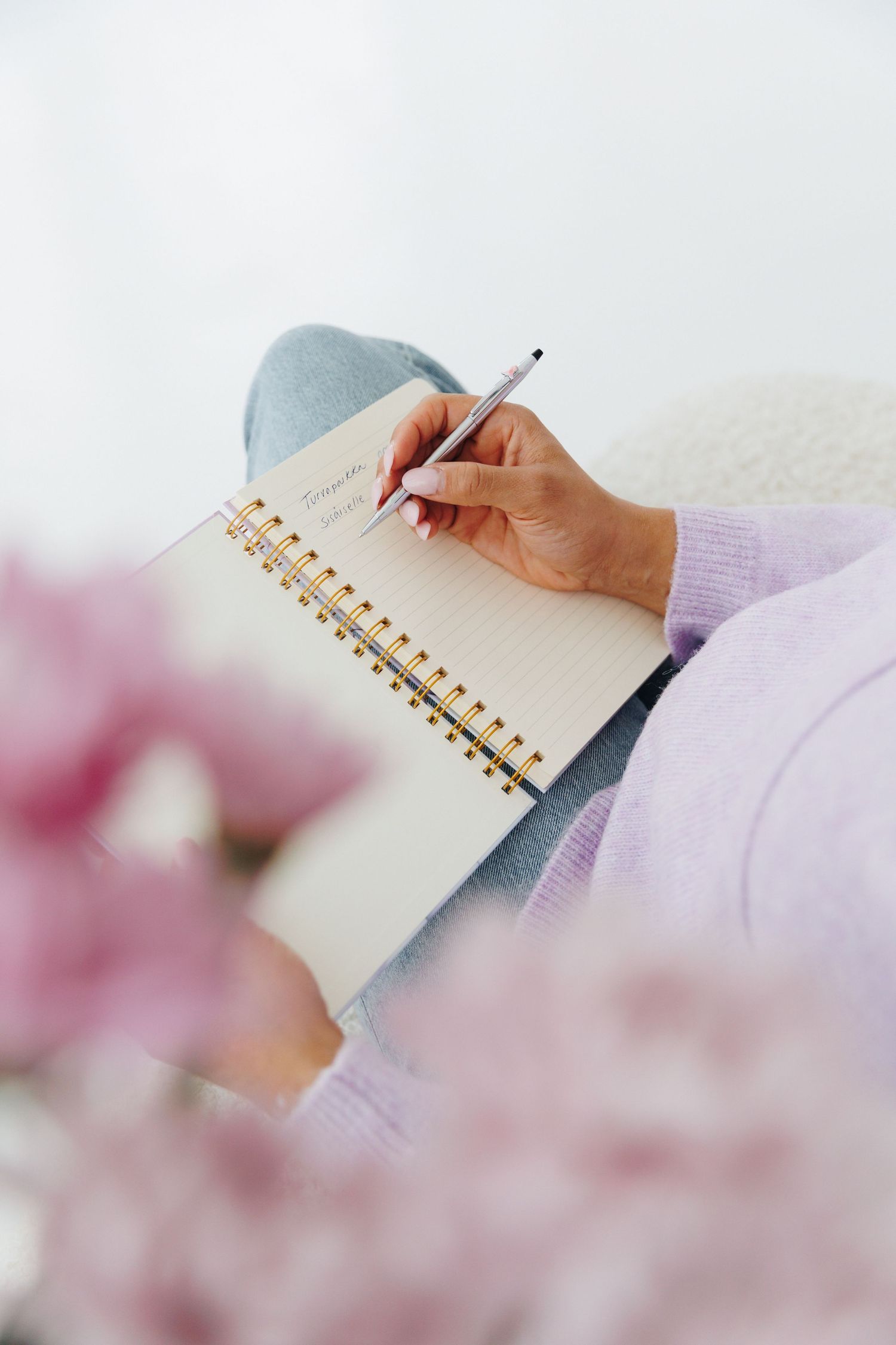 Close-up of a woman in a lavender sweater writing in a spiral notebook with a silver pen.