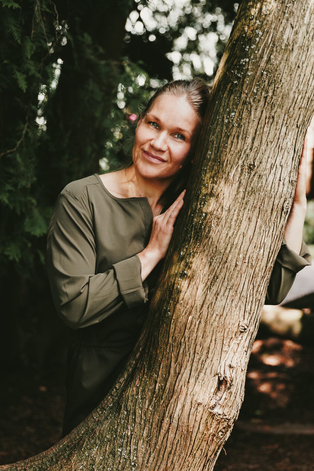 Smiling woman in olive top leaning against a large tree trunk in a lush forest setting.