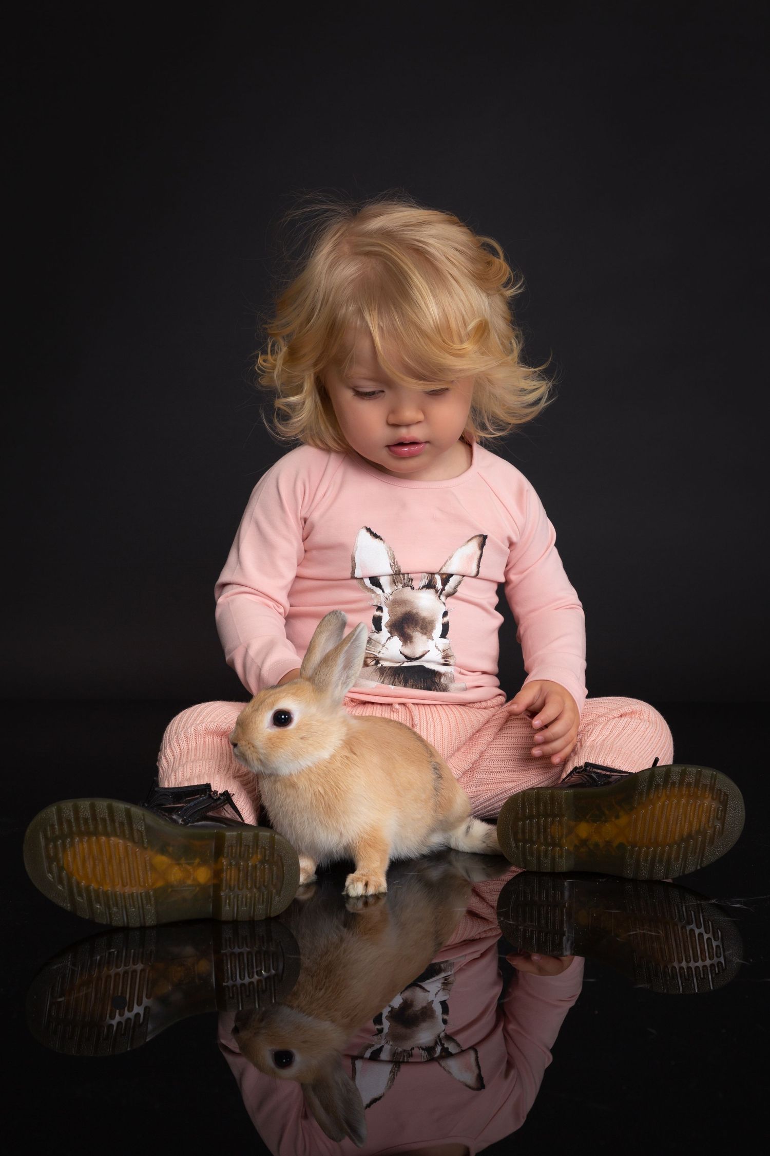 Toddler in pink outfit with curly blonde hair sits cross-legged on reflective floor with a small tan bunny rabbit.