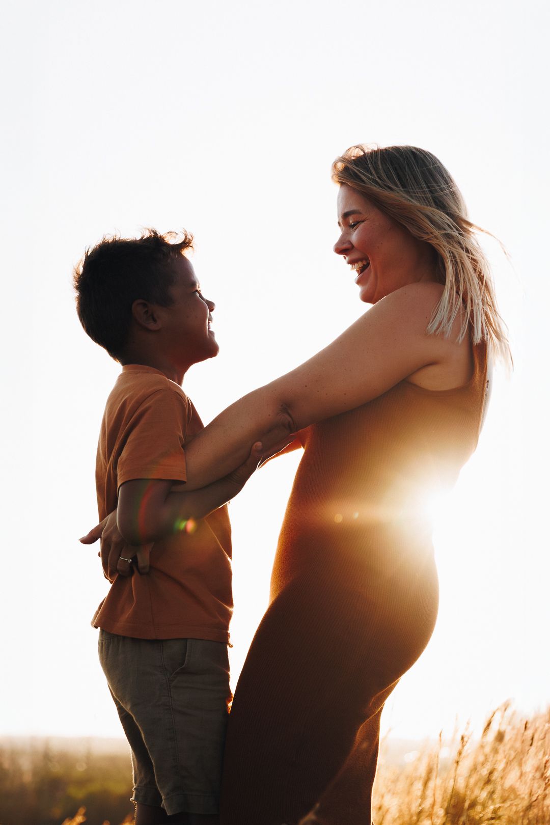 A silhouetted mother and child share a joyful moment in a sunlit field at sunset as sun rays stream through grass.