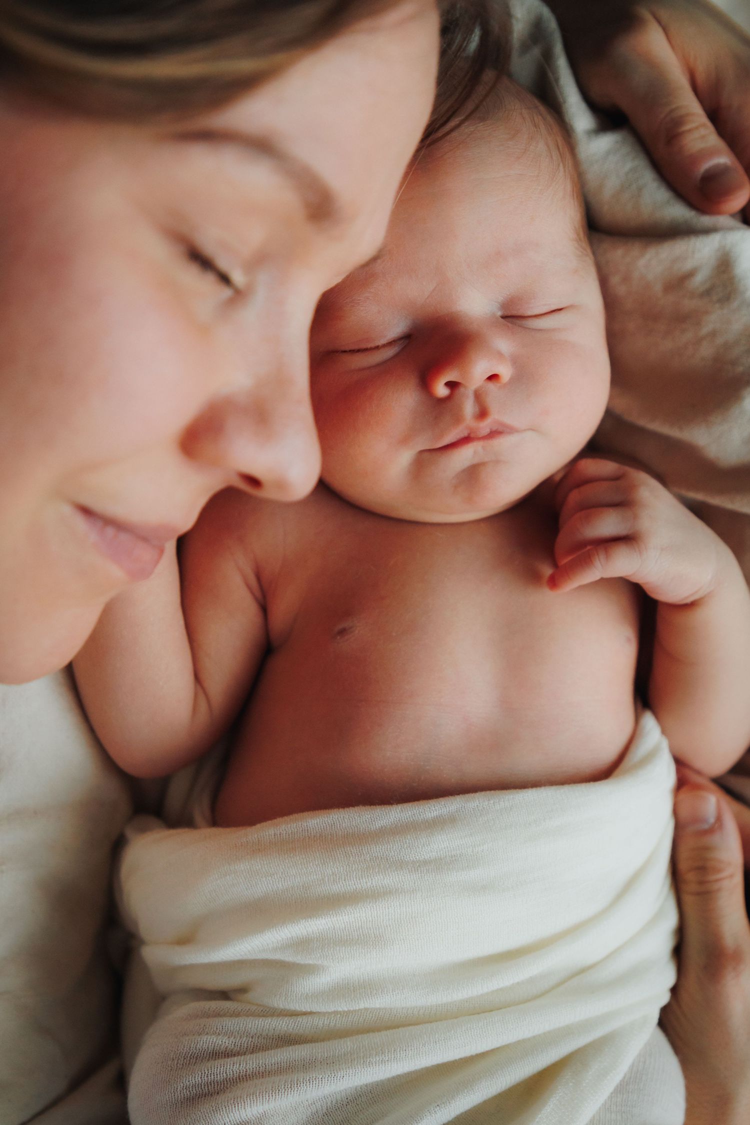 A tender moment captures parent and newborn baby sleeping peacefully together, wrapped in soft white fabric.