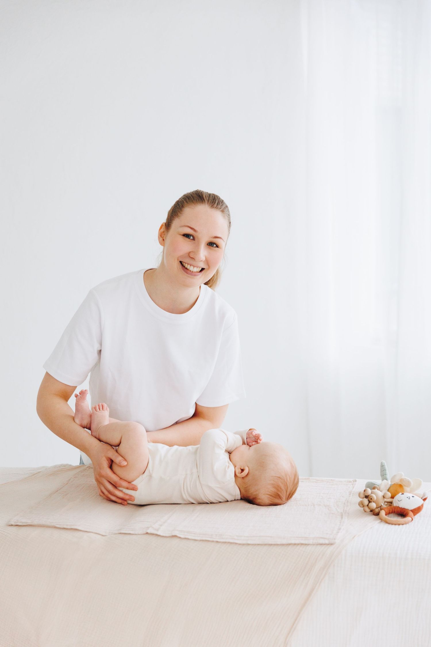 Smiling mother in white t-shirt doing baby massage on infant lying on soft blanket in bright white room.