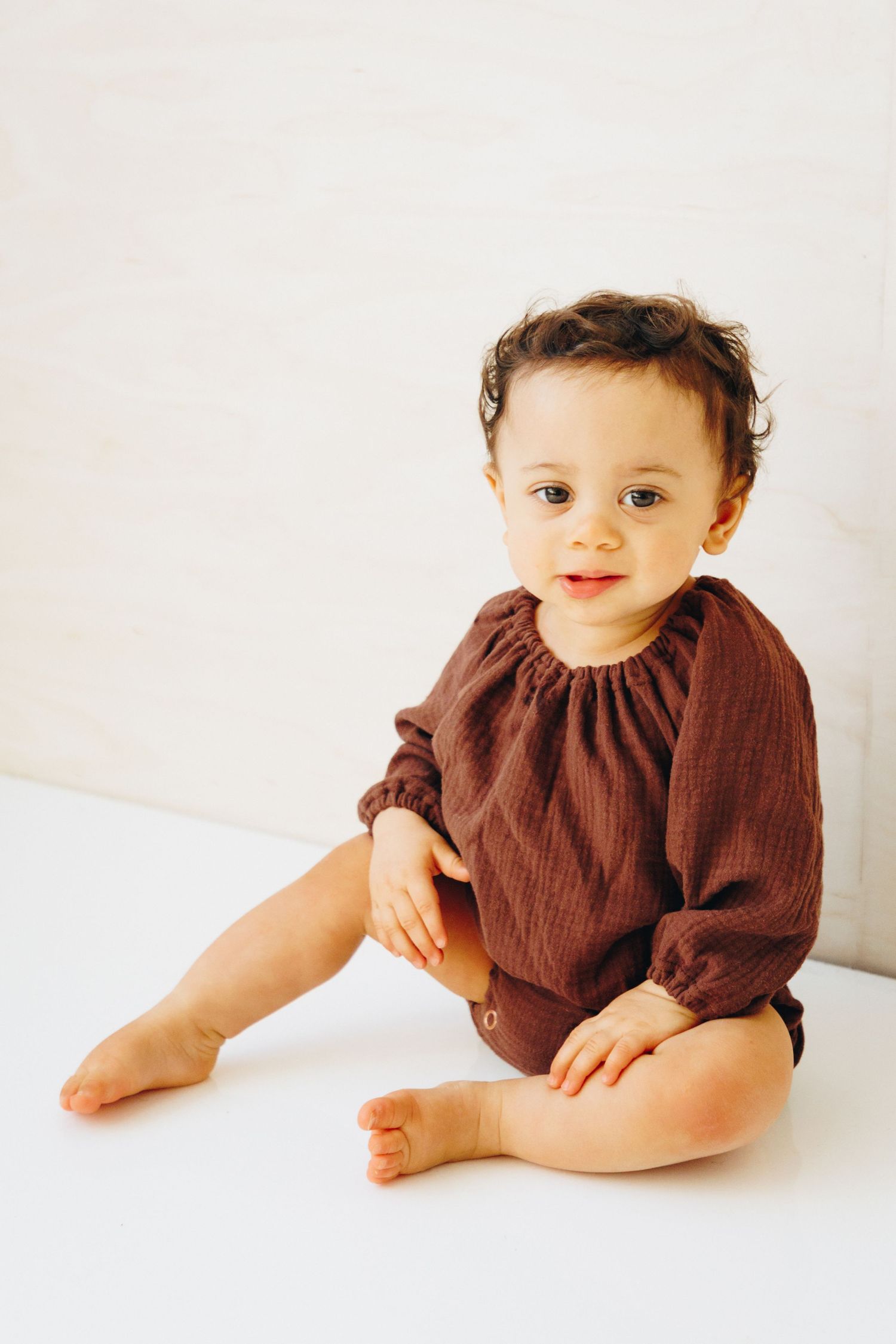 Cute baby in brown linen romper sitting on white surface, looking at camera with big eyes.
