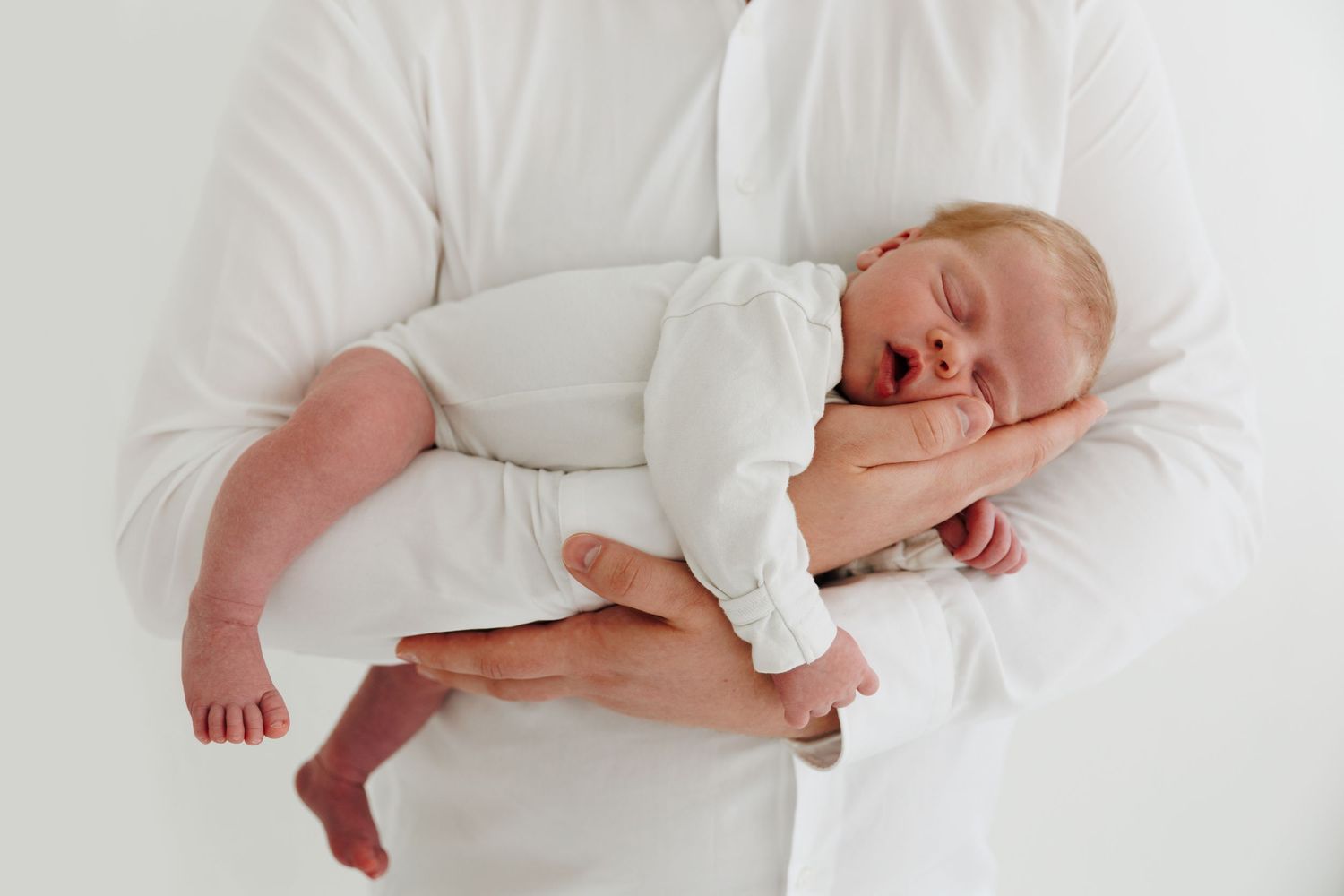 Adult in white shirt cradling a sleeping newborn baby dressed in white against a light background.
