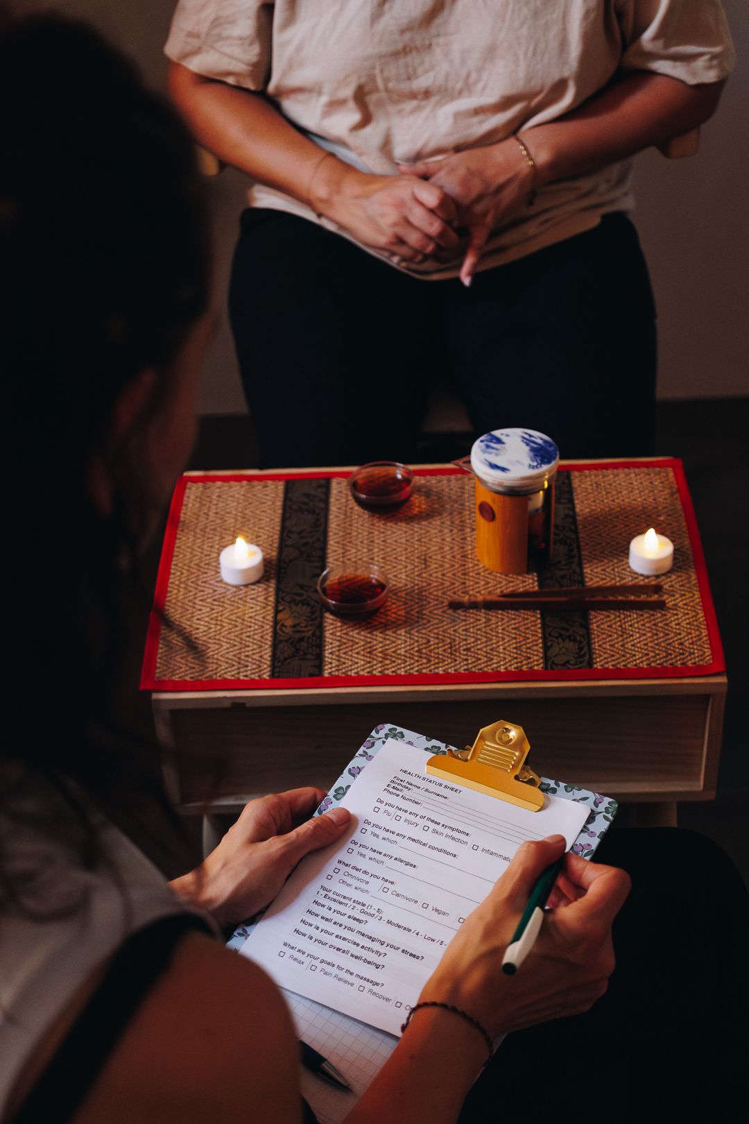 A therapist reviews a wellness intake form on a clipboard near a serene tea ceremony setup with candles and herbal tea.