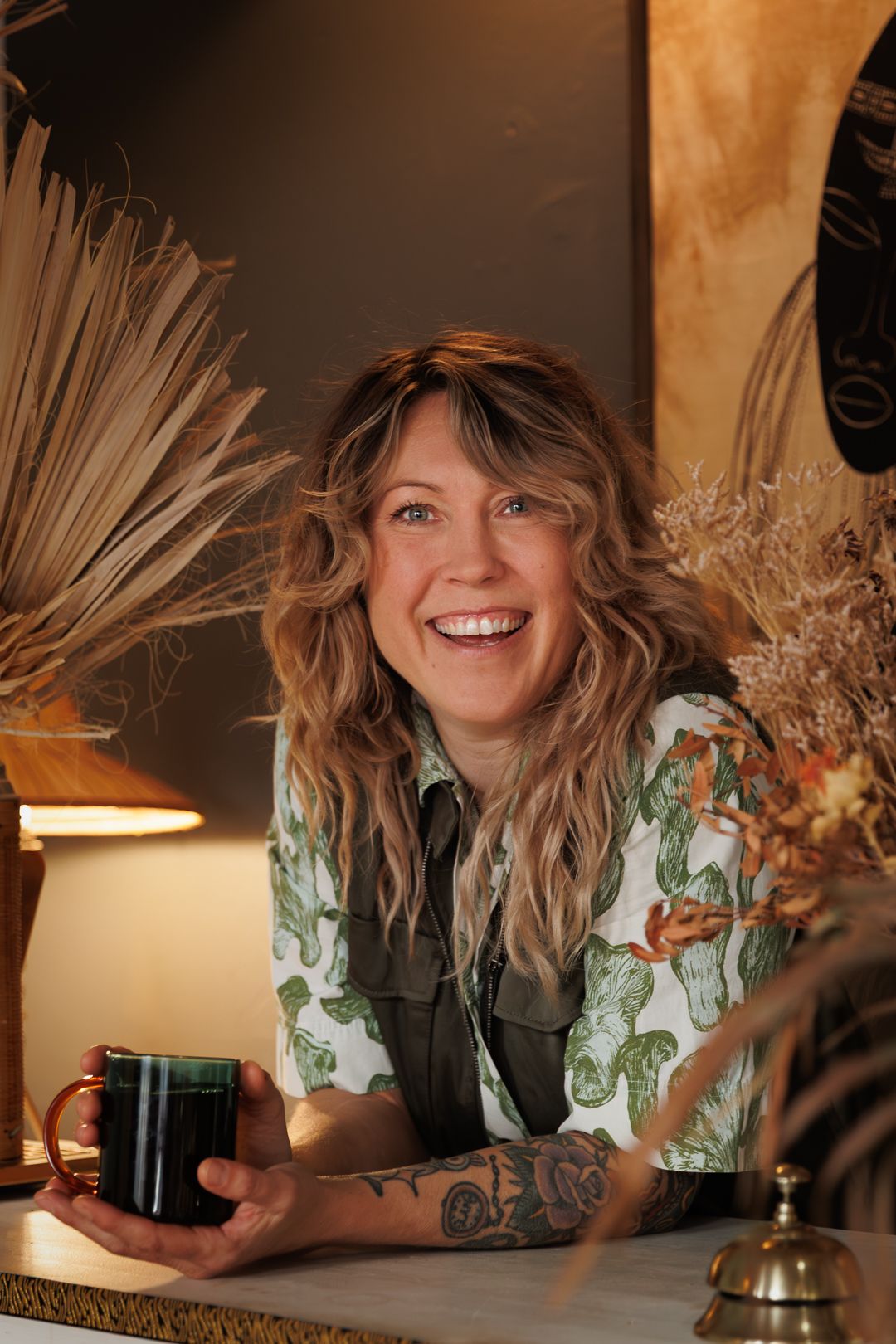 Smiling woman with wavy hair and tattoos holds a dark mug, seated near a lamp and dried floral decor.